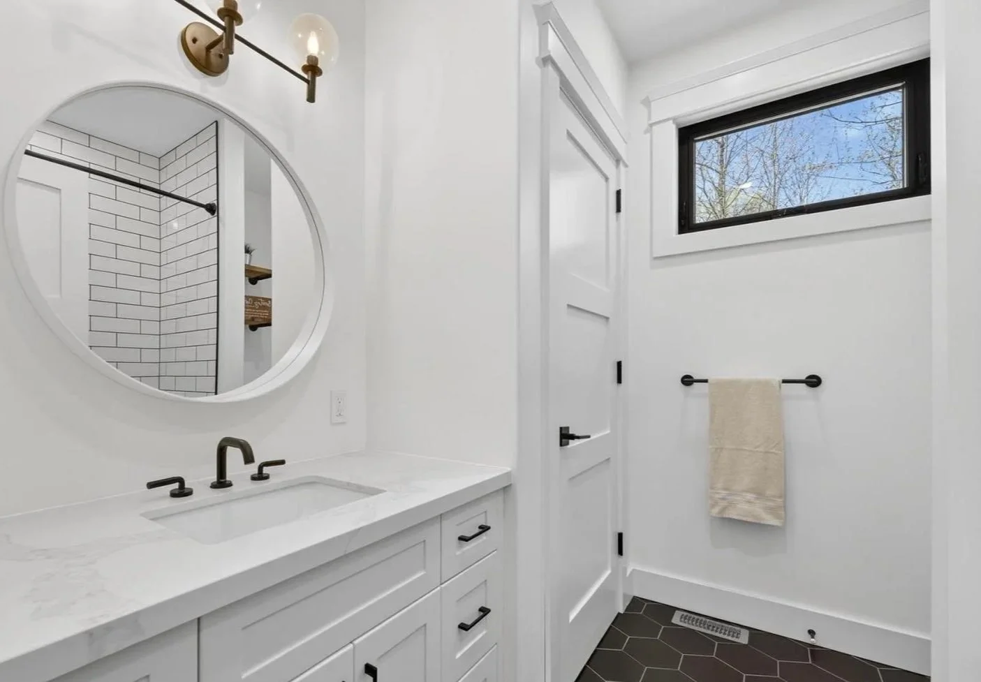 Modern white bathroom with a vanity, oval mirror, black hardware, tile shower seen through mirror, black-framed window, and beige towel on a black towel rod.