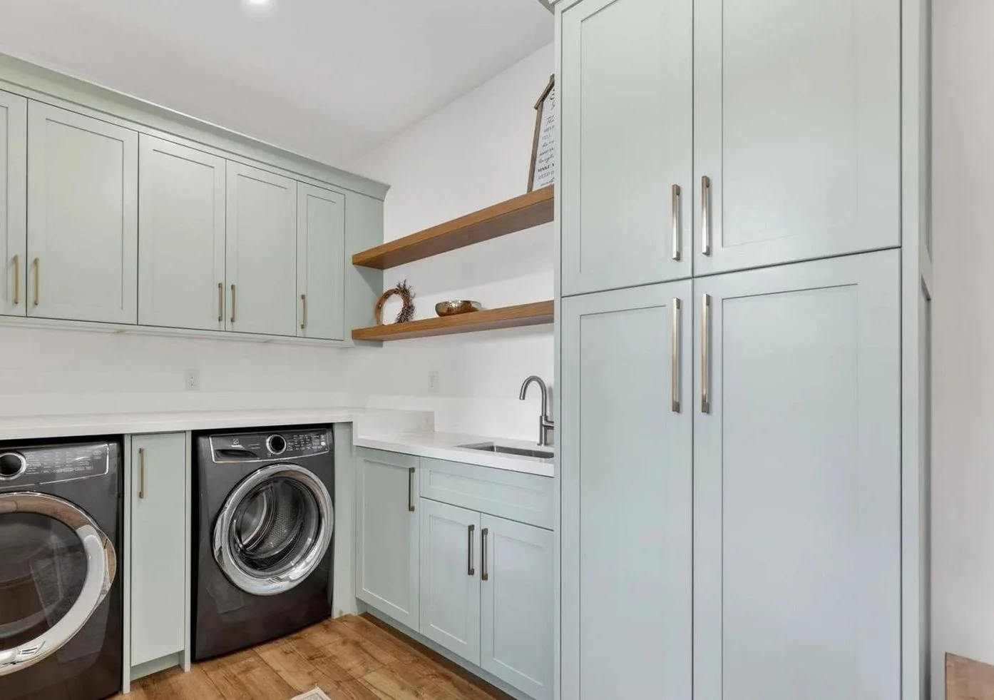 Laundry room with light green cabinets, a white countertop, a stainless steel sink with a faucet, open wooden shelves with decorative items, a front-loading washing machine, a front-loading dryer, and hardwood floors.