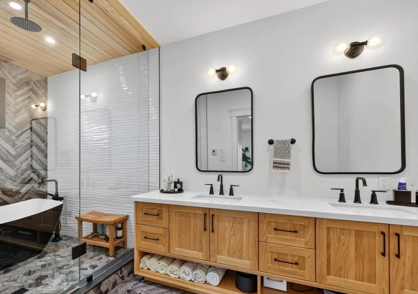 Modern bathroom with dual black-framed mirrors above a wooden vanity with two sinks, black fixtures, and a white countertop. Shower area with textured glass wall, a freestanding bathtub, and stylish wood and tile decor.