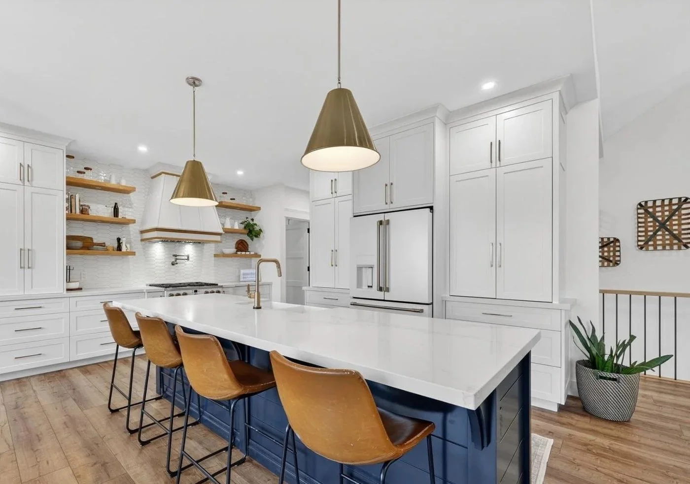 Modern kitchen with white cabinets, a large white island with a dark blue base, six brown bar stools, gold pendant lights, open wooden shelves, and hardwood floors.