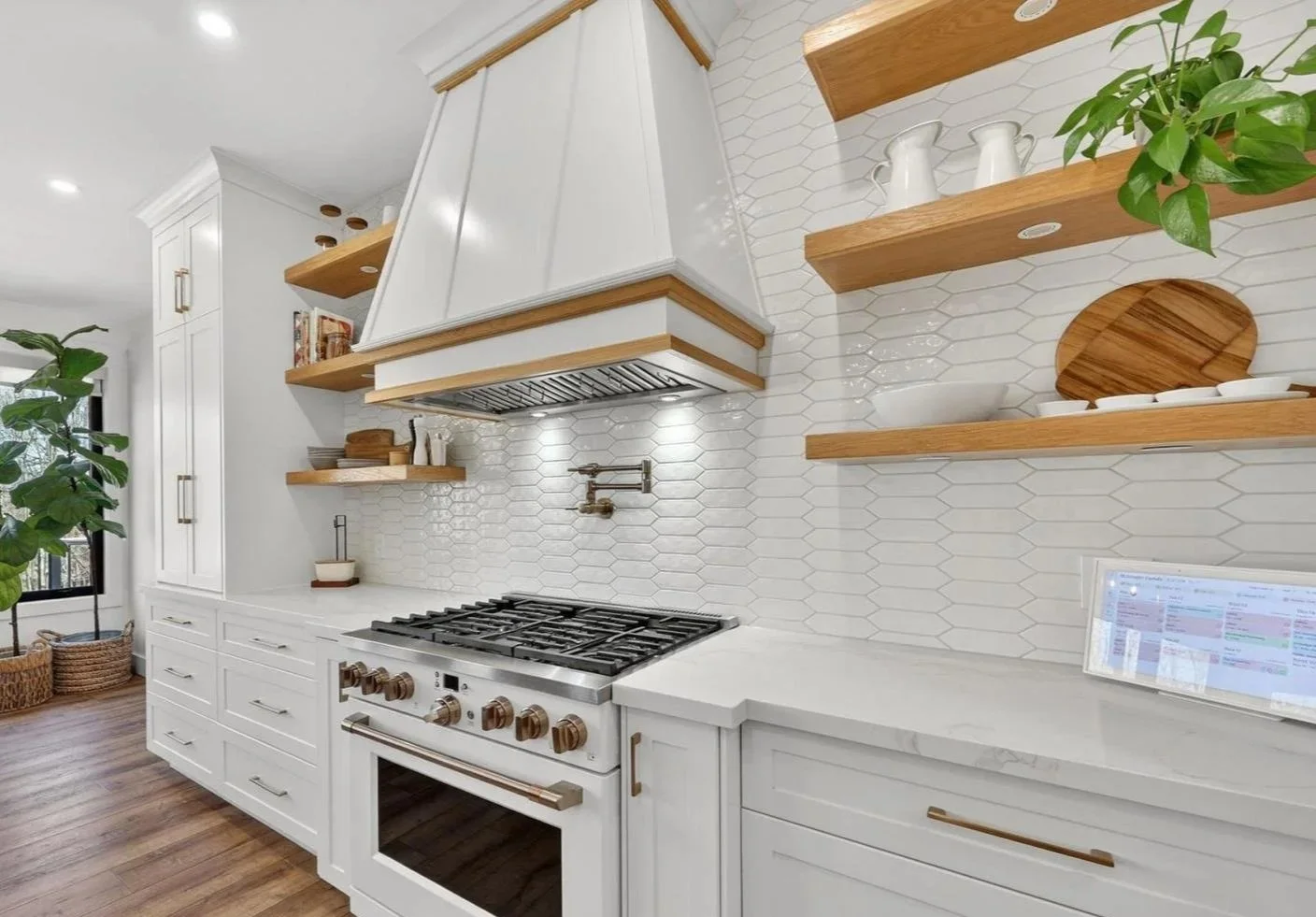 Modern kitchen with white cabinets, wooden open shelves, a white range hood, gas stove, white hexagonal tile backsplash, and a laptop on the white countertop.