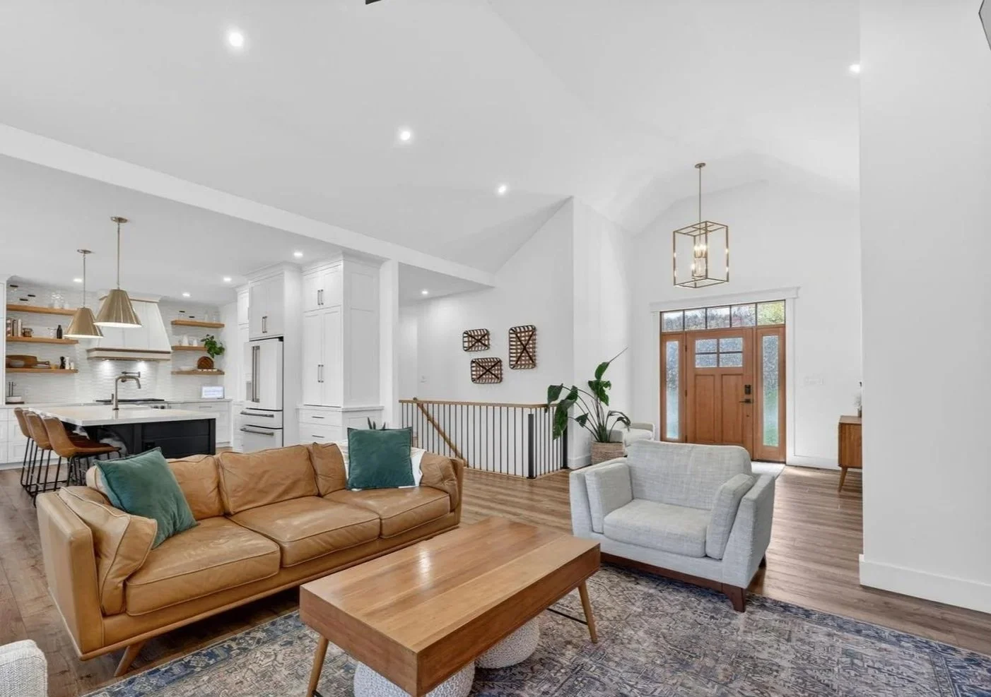 Living room and kitchen area with white walls, hardwood floors, a brown leather sofa with teal pillows, a light gray armchair, a wooden coffee table, and a front door with glass panels and a wooden frame. Modern lighting fixtures and decorative wall 
