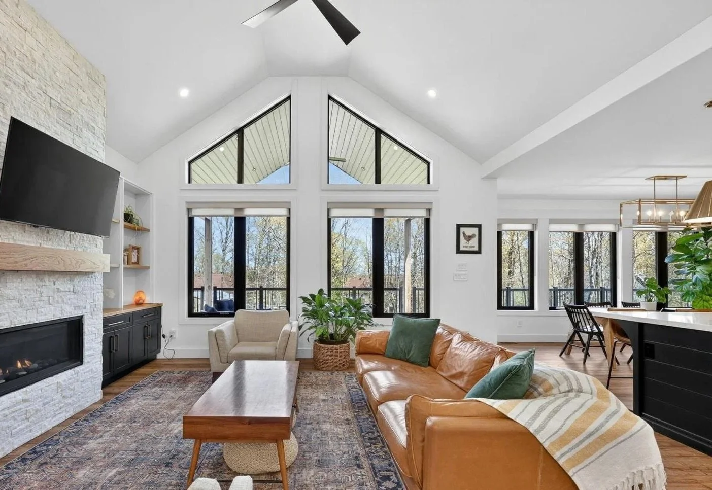 Living room with white walls and high vaulted ceiling, large windows, and a ceiling fan. Features a leather sofa with cushions, a beige armchair, a wooden coffee table, a patterned rug, a white fireplace with a black surround, built-in shelves, and h