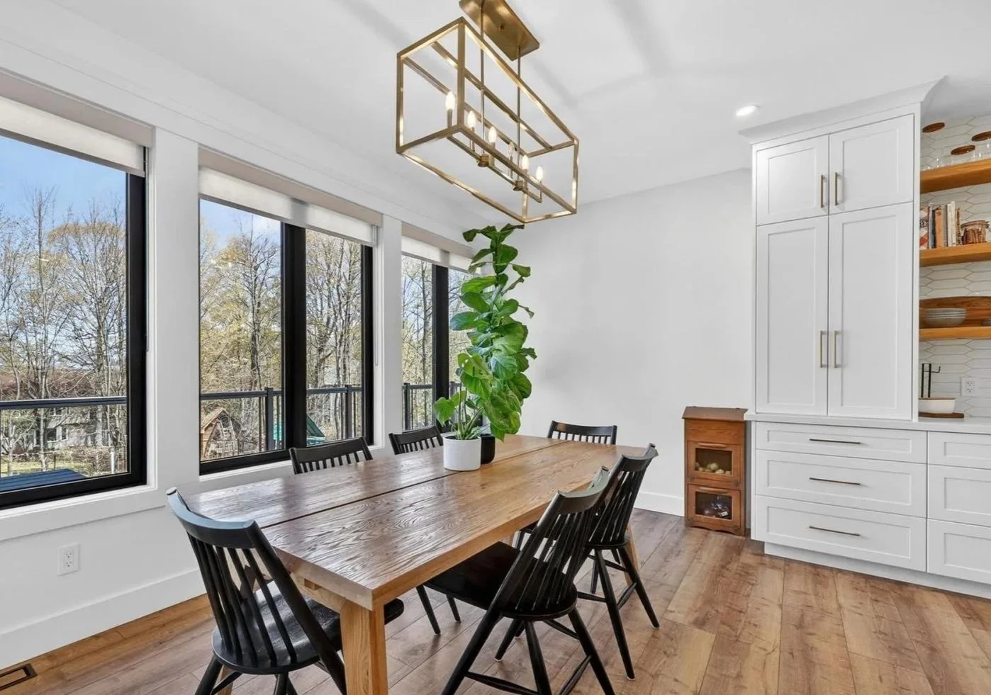 A dining room with a wooden table, six black chairs, a large green potted plant, four windows, and a white cabinet with open shelves and drawers. There is a modern light fixture hanging from the ceiling.