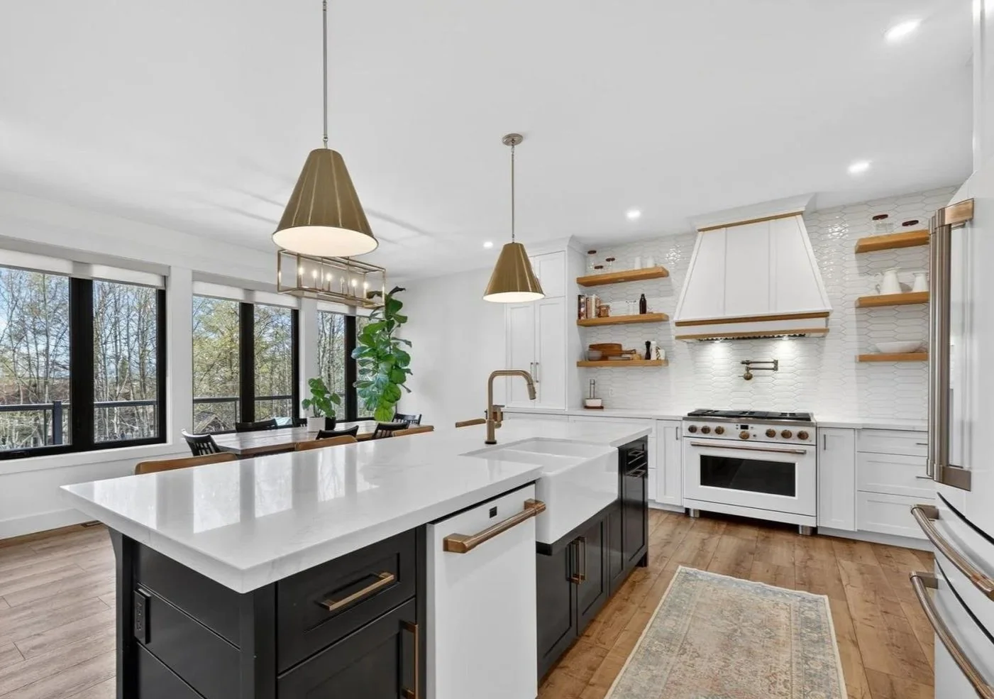 Modern kitchen with white cabinets, wood open shelves, black and white island, gold hardware, and large windows with trees outside.