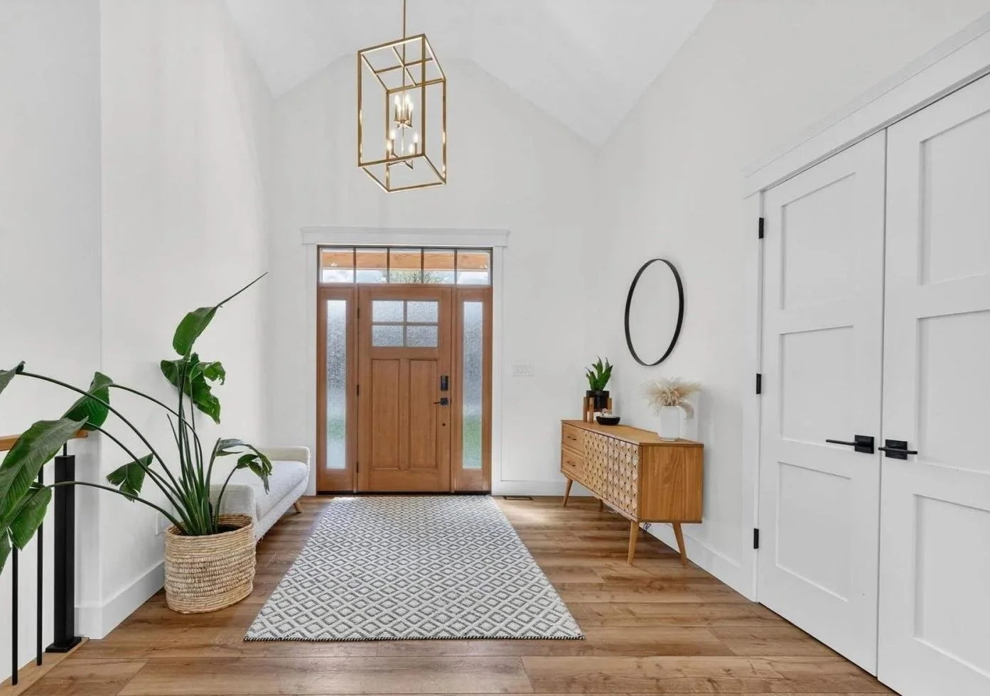 Modern entryway with wooden front door, potted plants, a patterned rug, white walls, and a wooden sideboard with decor.