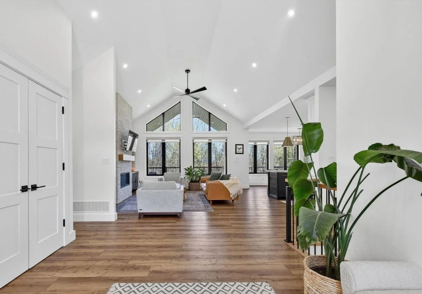 Spacious living room with vaulted ceiling, large windows, white walls, hardwood floors, modern furniture, and indoor plants.