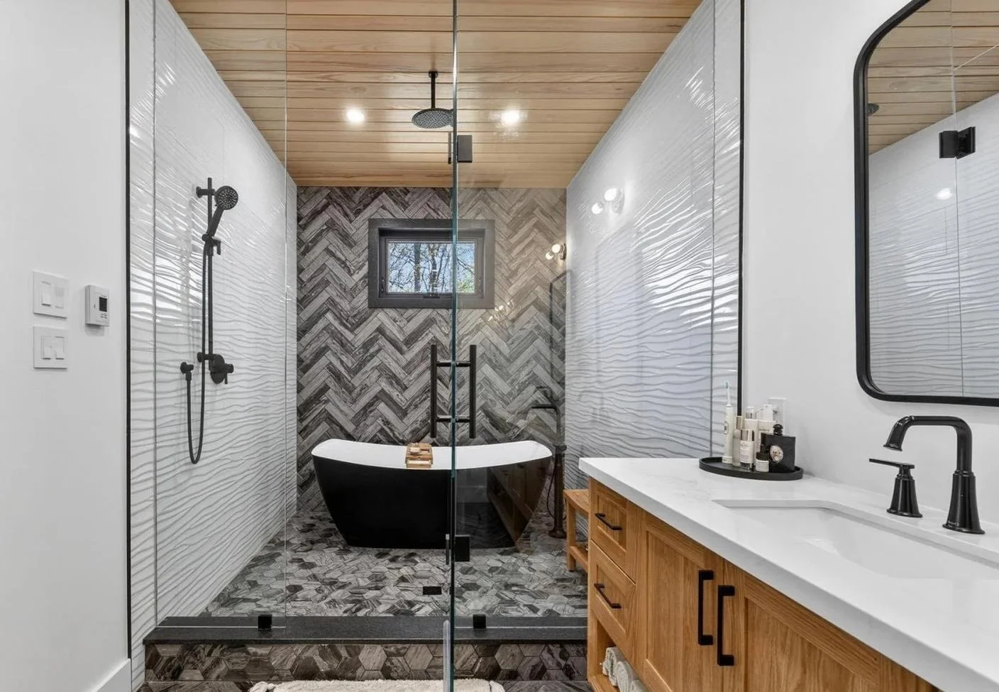 Modern bathroom with glass-enclosed shower and wooden vanity with black fixtures, a freestanding bathtub, patterned tile wall, and a window.