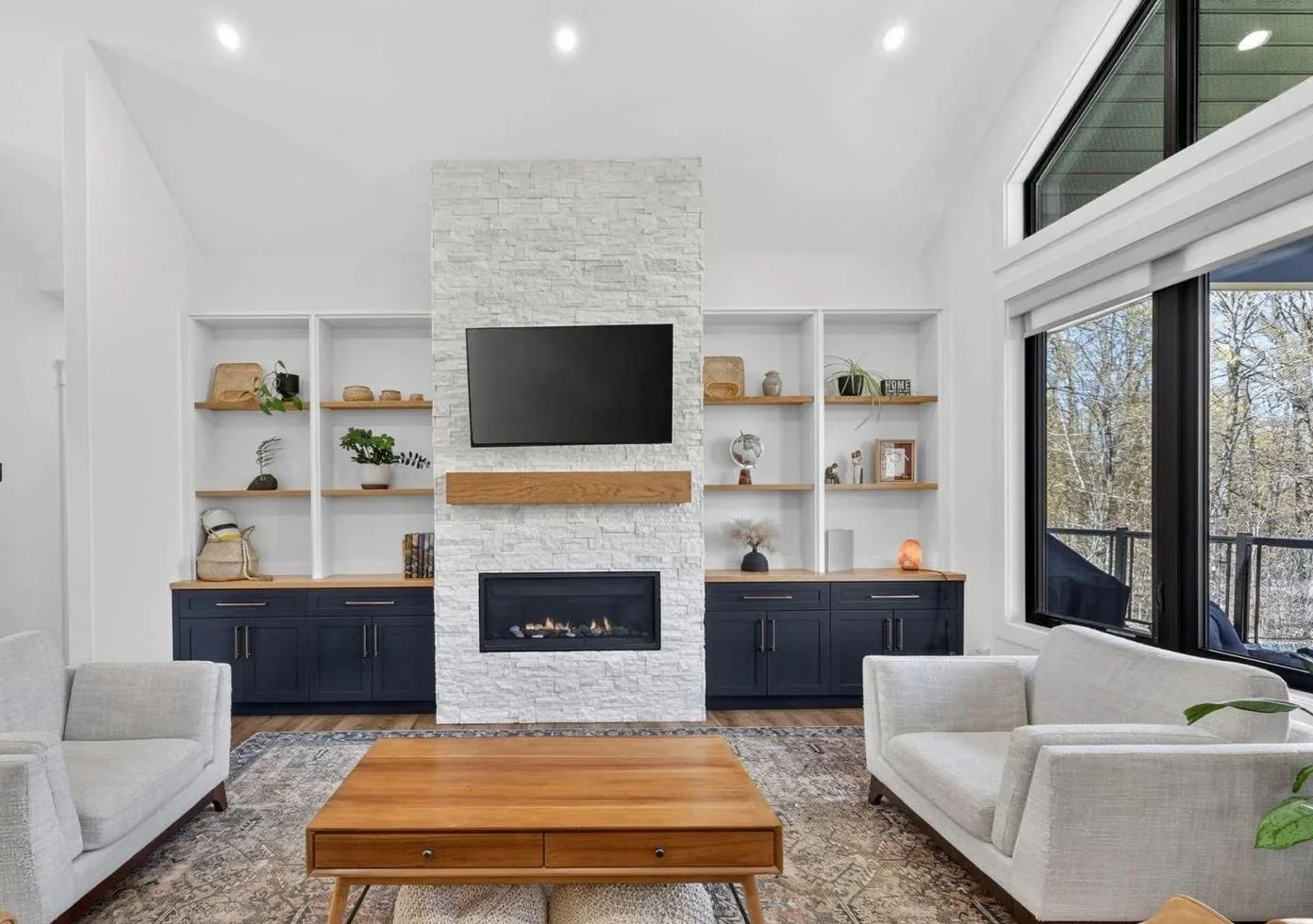 Living room with white walls, large windows, and a modern fireplace with a TV mounted above. Built-in shelves with decor, a wooden coffee table, and light-colored sofas.
