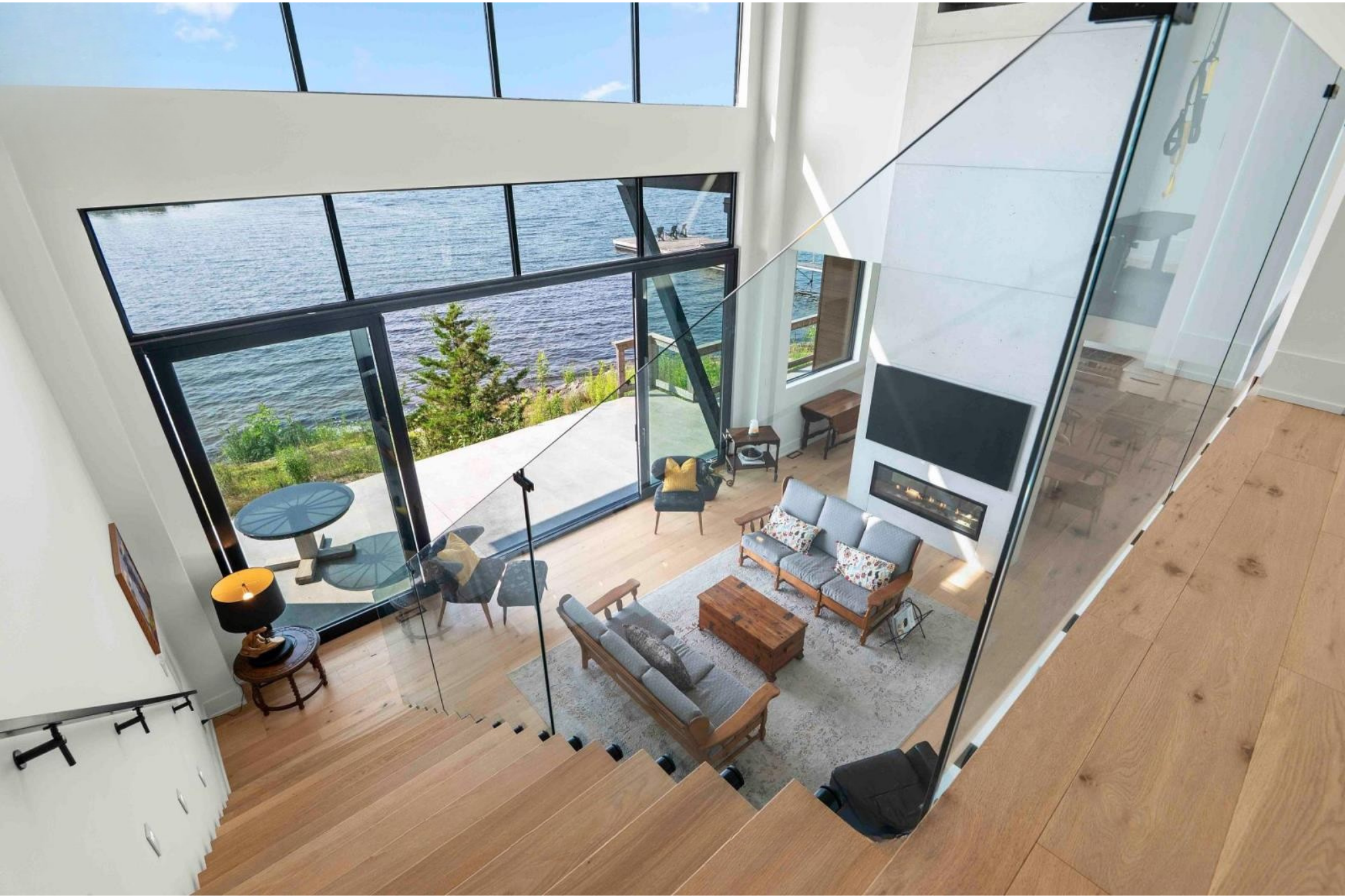 Interior view of a modern living room with floor-to-ceiling windows overlooking a body of water, featuring a wooden coffee table, gray sofas with patterned pillows, a blue armchair, and a fireplace. A staircase with wooden steps is visible in the for