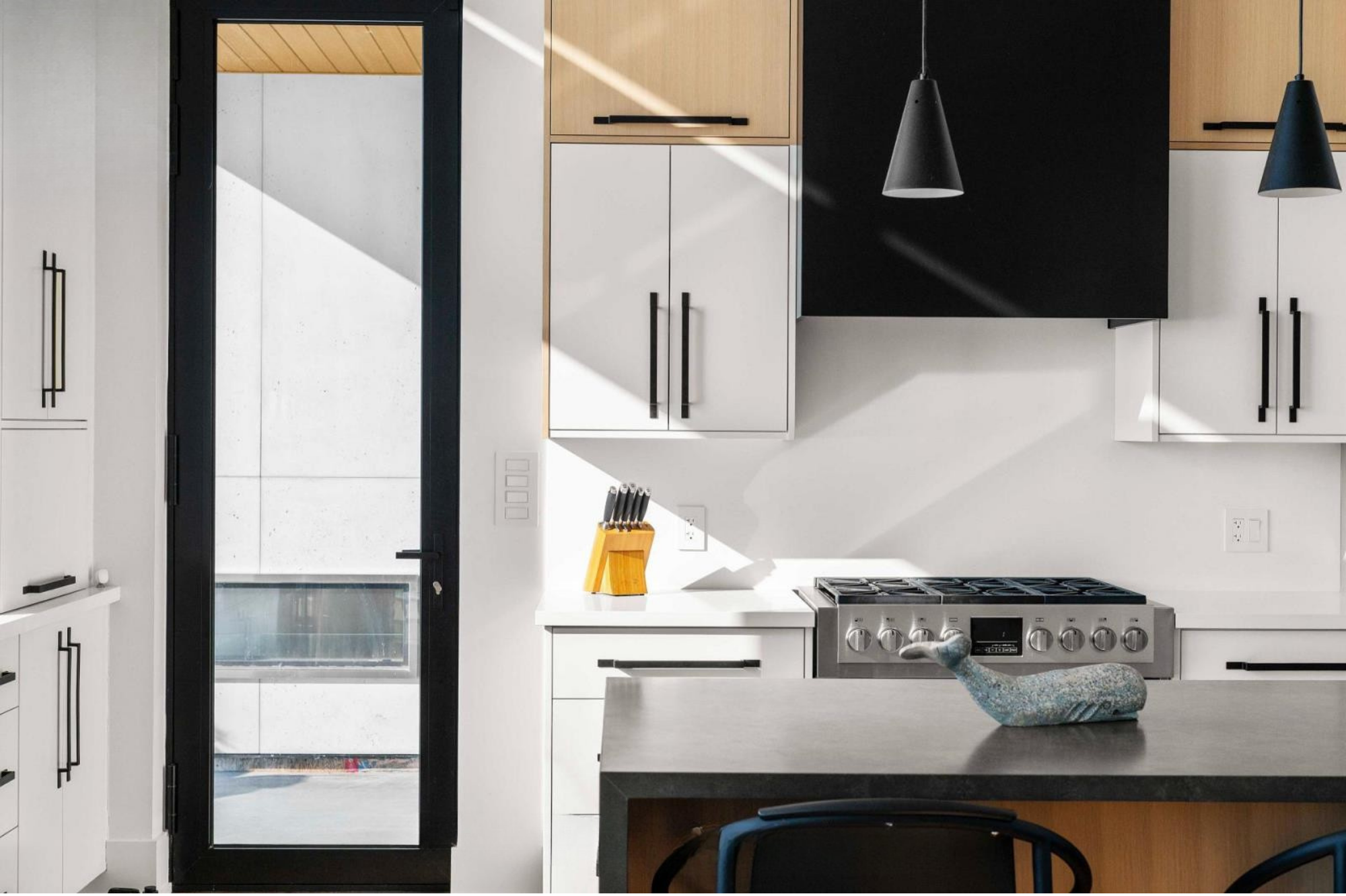 Modern kitchen with white and black cabinetry, a stainless steel stove, black pendant lights, a knife block on the white countertop, and a black door with a glass panel leading outside.