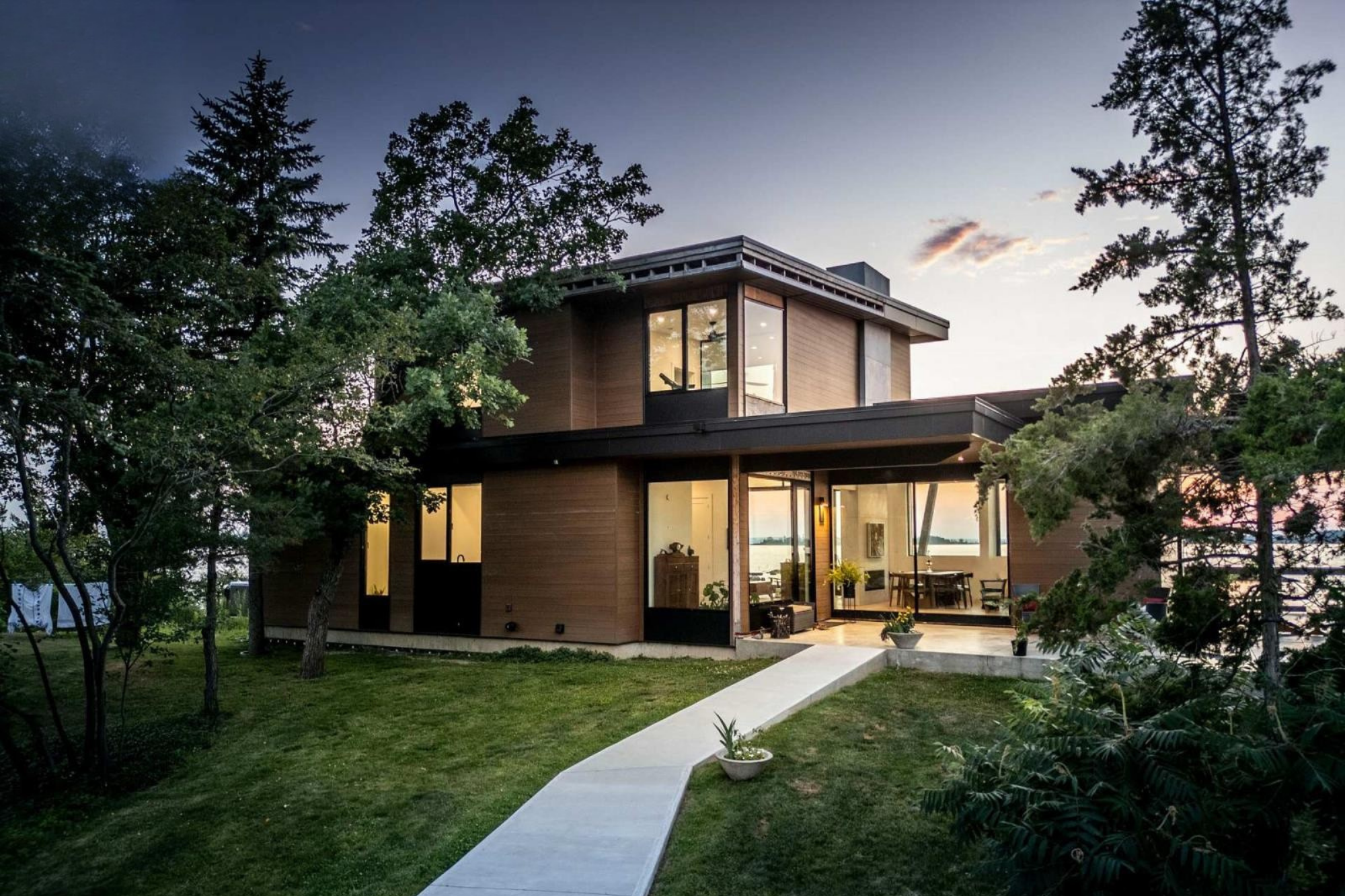 Modern two-story house with large glass windows, wooden exterior, and an open patio, surrounded by trees at dusk.