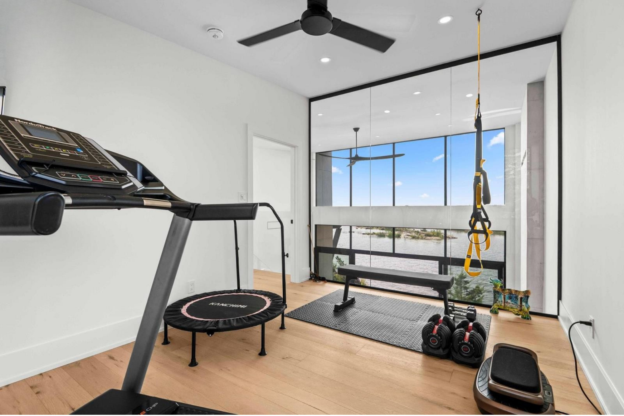 Home gym with treadmill, mini trampoline, dumbbells, punching bag, resistance band, and bench in front of large window showing blue sky and clouds.