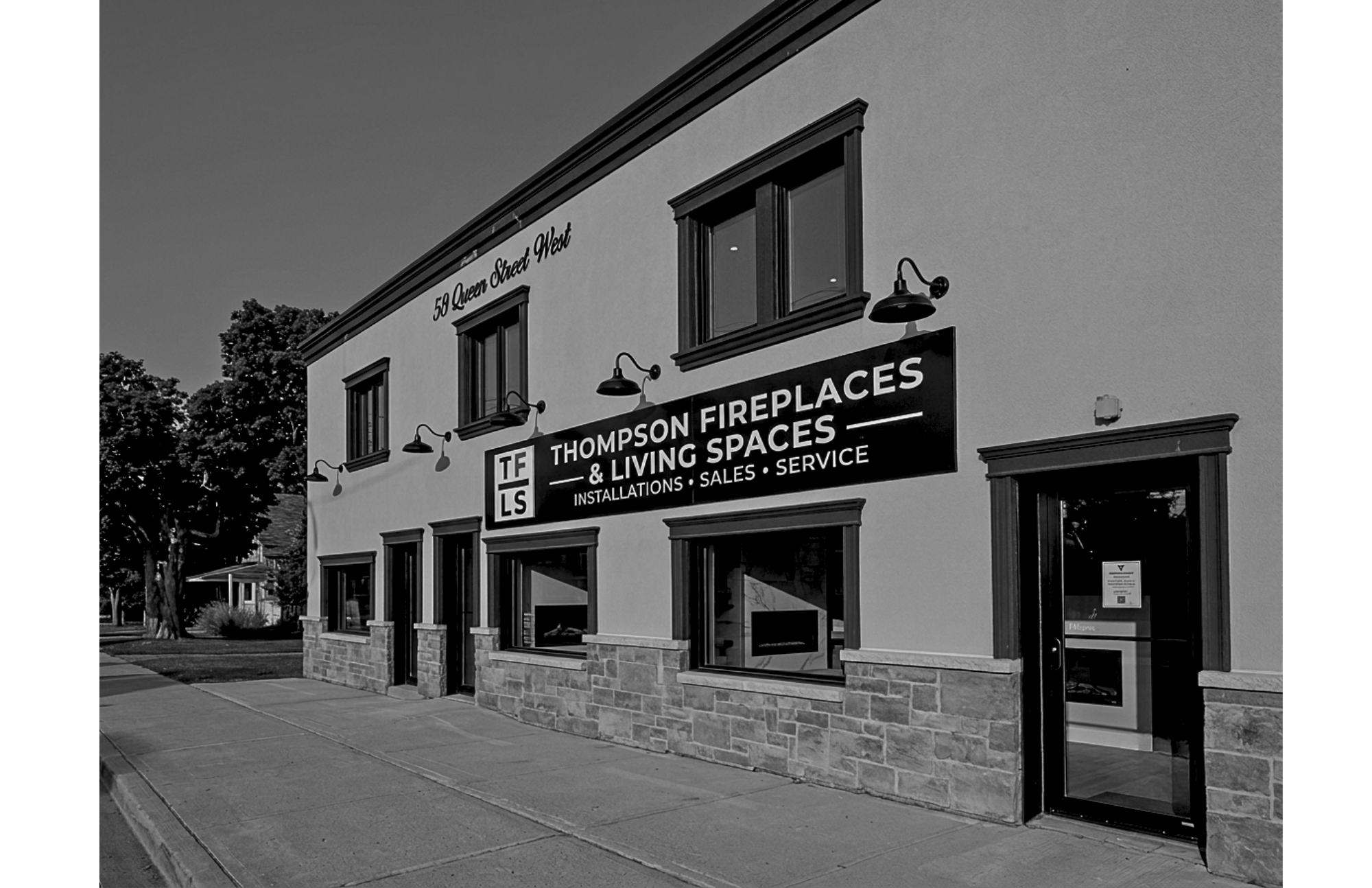 Exterior of a building with a sign for Thompson Fireplaces & Living Spaces, showing services like installations, sales, and service. The building has three windows on the upper floor with dark trim and exterior lighting fixtures. There is sidewalk and trees visible in the surrounding area.
