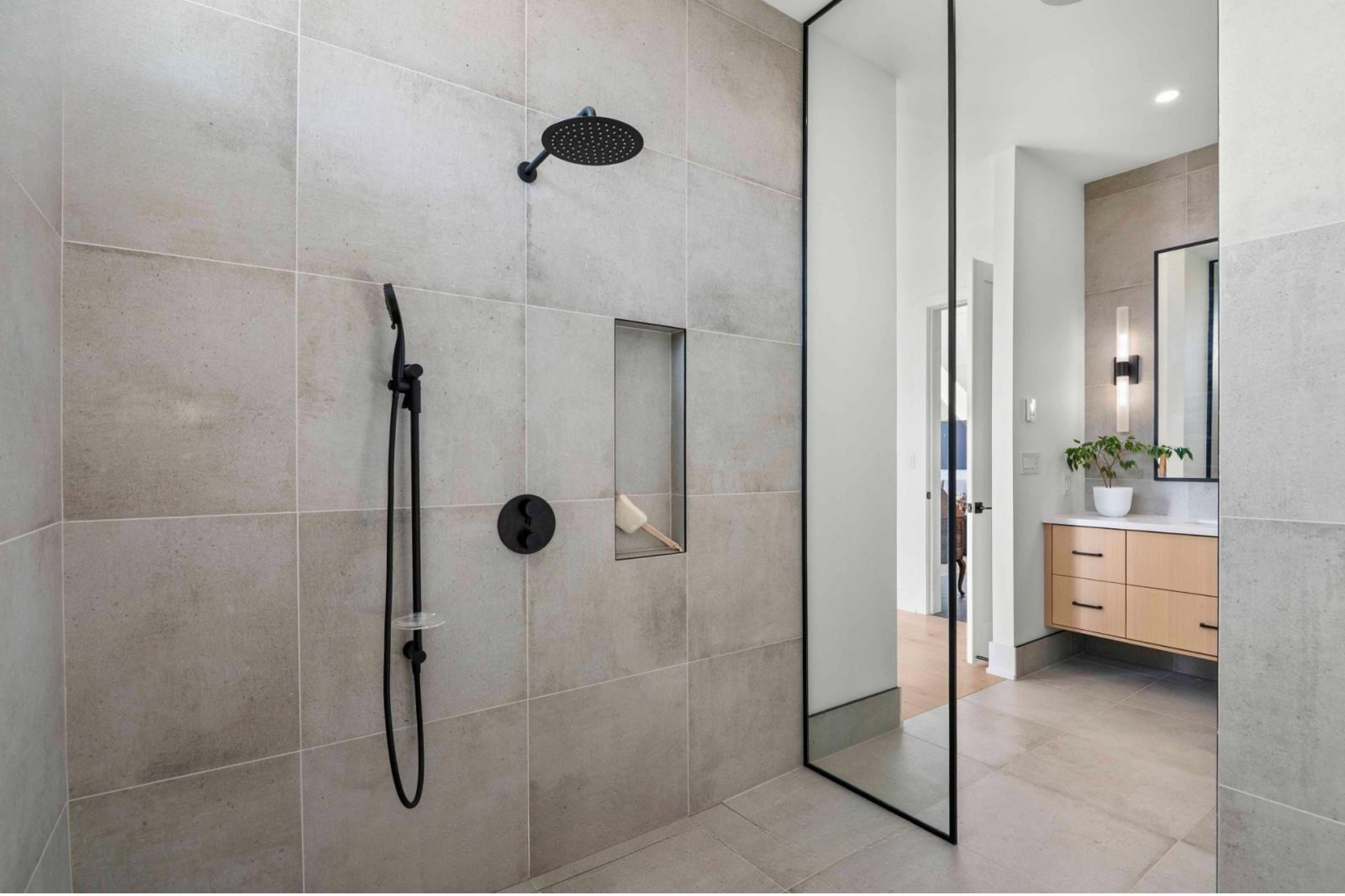 Modern bathroom with large tiled shower area, black shower fixtures, built-in shelf, and a large mirror reflecting a vanity with a white pot and green plant.