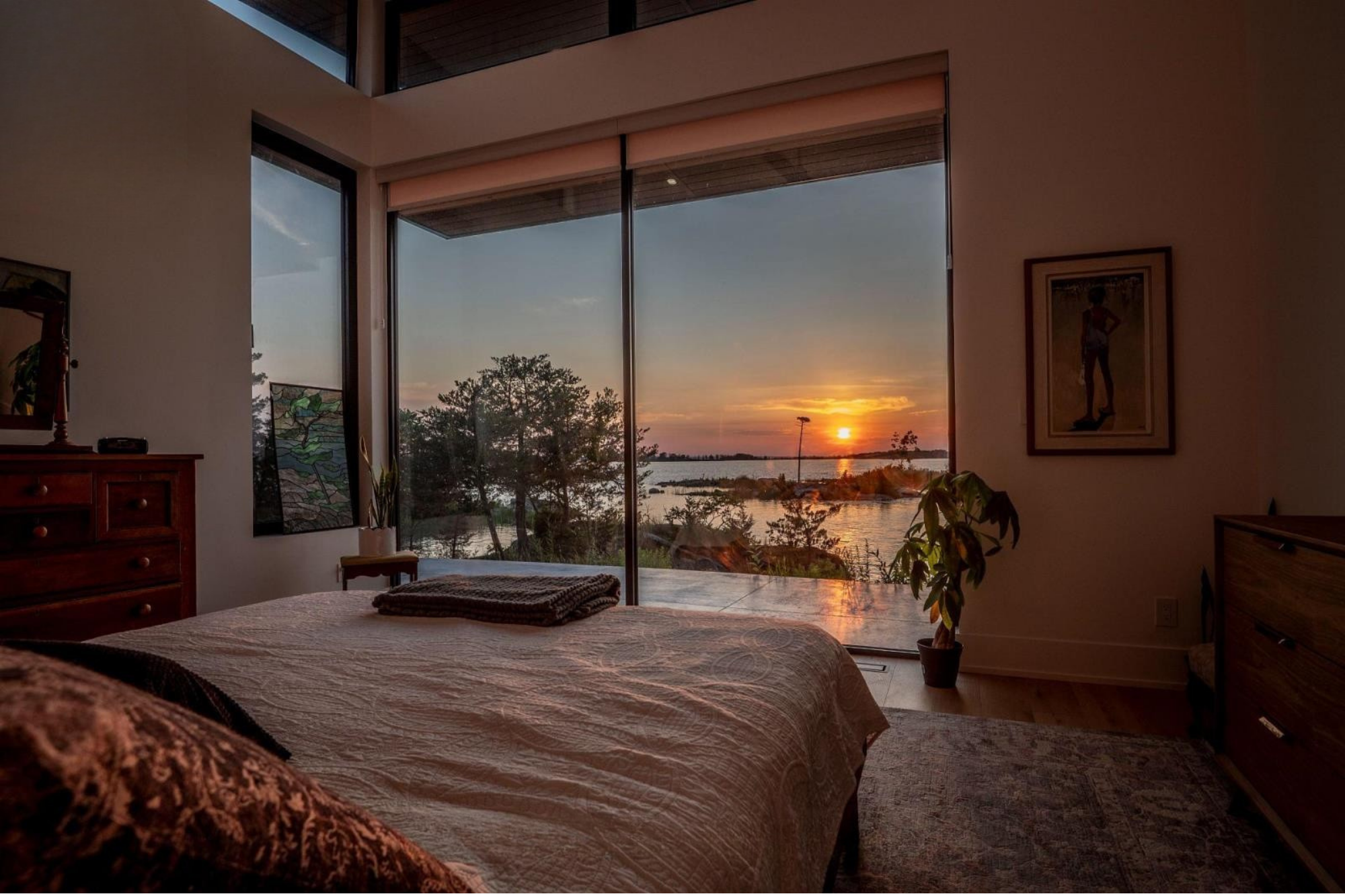 Interior of a bedroom with large window showing a sunset over a body of water and trees outside. The room has a wooden dresser, framed pictures, a potted plant, and a bed with a beige quilt, with a folded blanket at the foot.