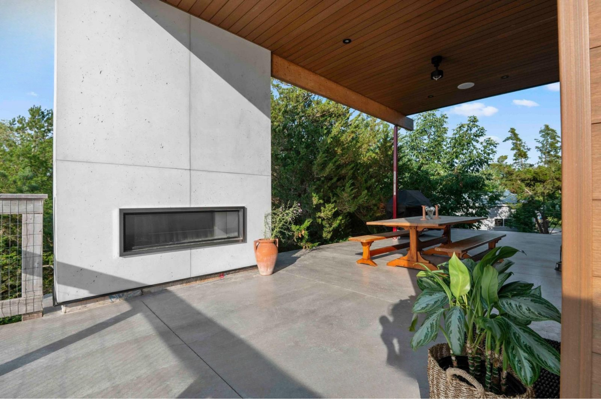 Modern outdoor patio with a concrete floor, a wooden ceiling, and a wooden picnic table with benches. There are green plants in woven baskets on the floor, and a background of trees and a blue sky with some clouds.