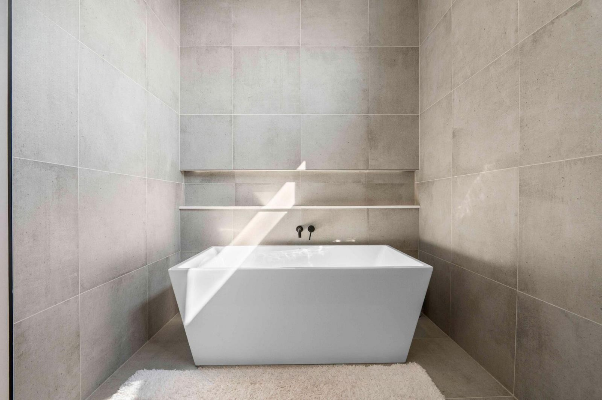 A modern bathroom with a white rectangular bathtub against gray tiled walls, a white fluffy rug on the floor, and a shelf above the bathtub.