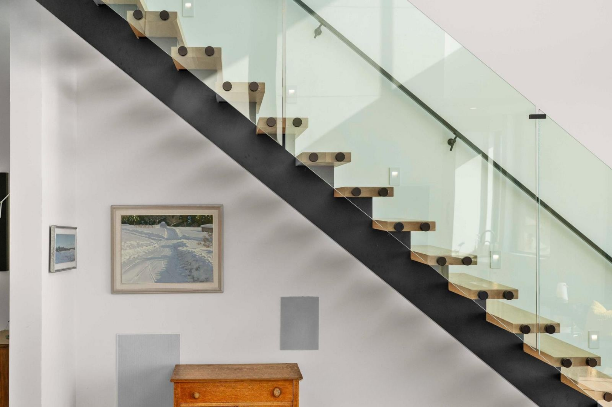 Interior view of a modern staircase with wooden steps, black edge, and glass railing, in a well-lit room with framed artwork and a wooden furniture piece on the wall.