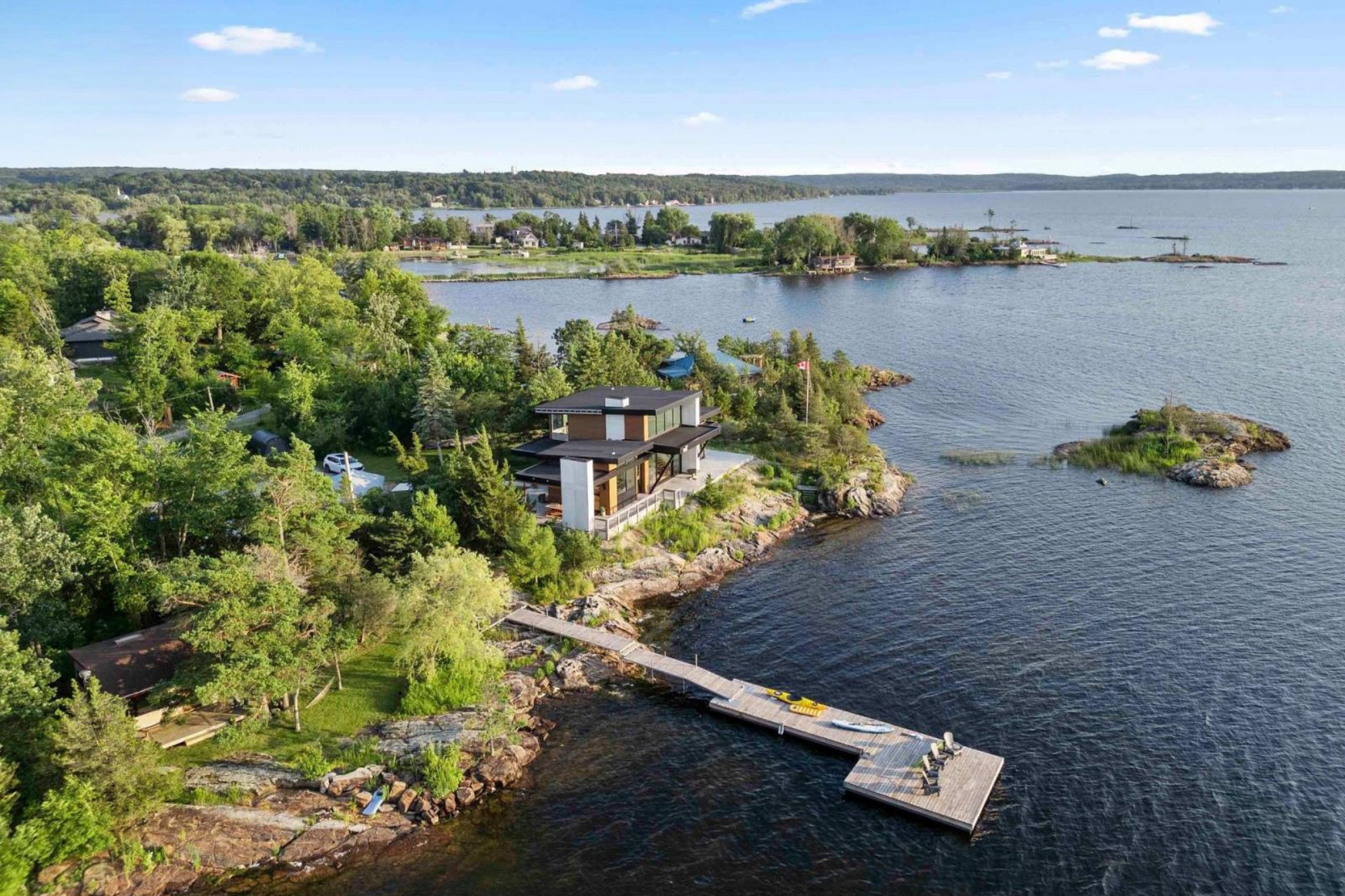 Aerial view of a modern house with flat roof on rocky shoreline, surrounded by dense green trees, with a dock extending into a large body of water. In the background, there are more houses and a vast lake under a blue sky with a few clouds.