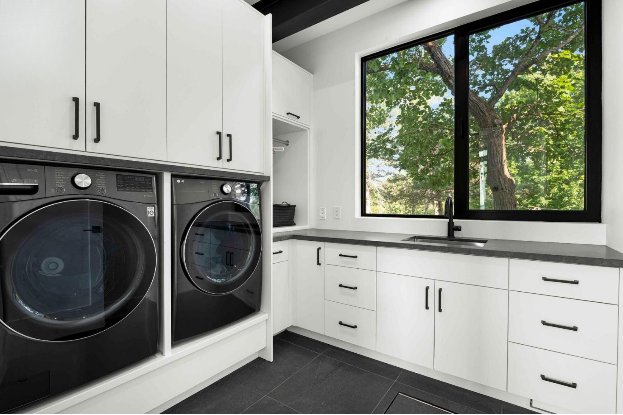 Modern laundry room with white cabinetry, black cabinet handles, a large window showing green trees outside, and black front-loading washing machines.