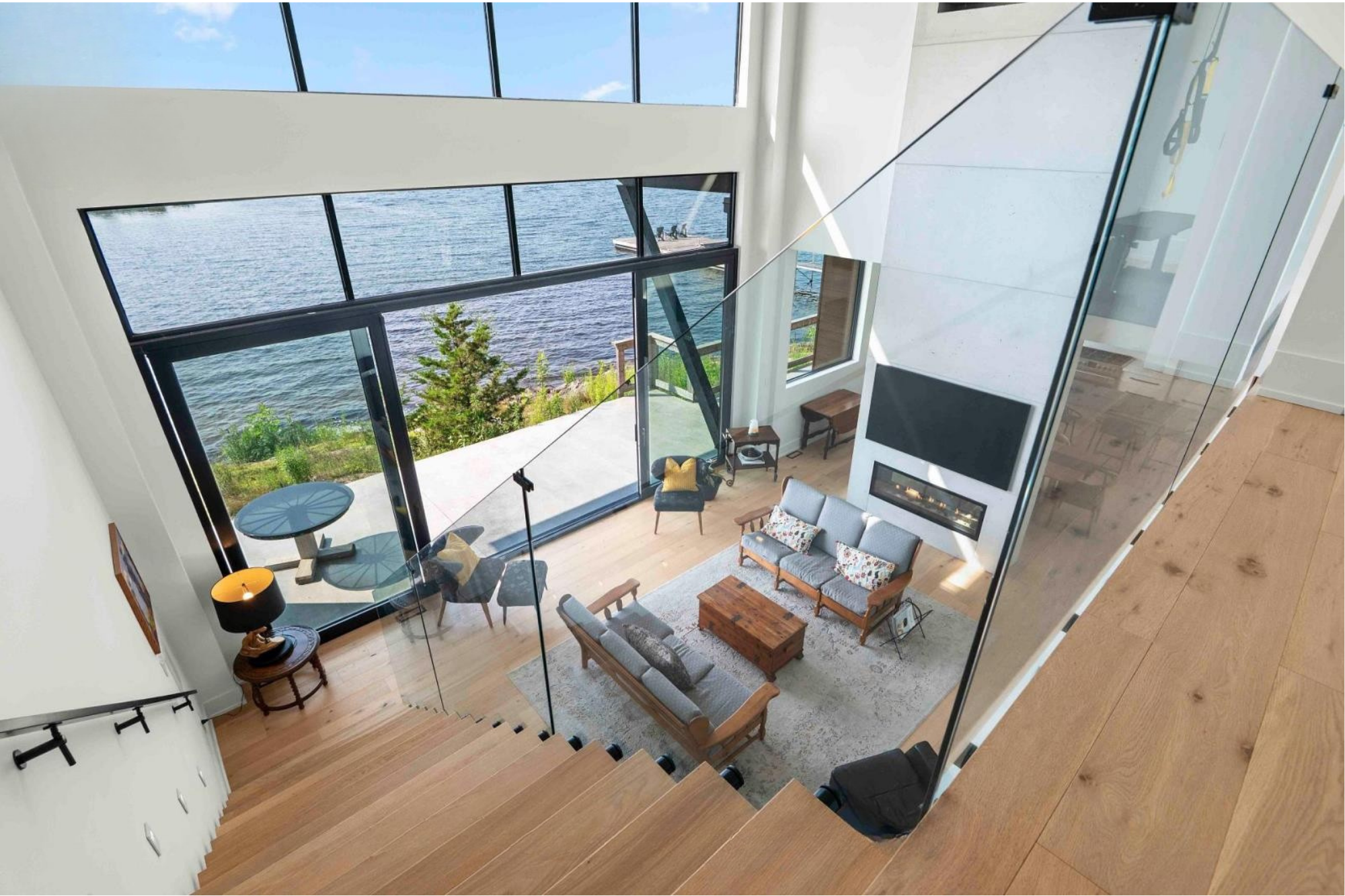 Modern living room with floor-to-ceiling windows overlooking a body of water, featuring furniture including a sofa, armchair, coffee table, dining table with chairs, and a fireplace.