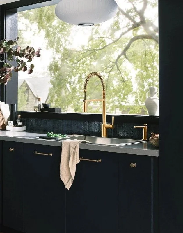Modern kitchen with black cabinets, white countertop, gold sink faucet, and a large window showing green trees outside.