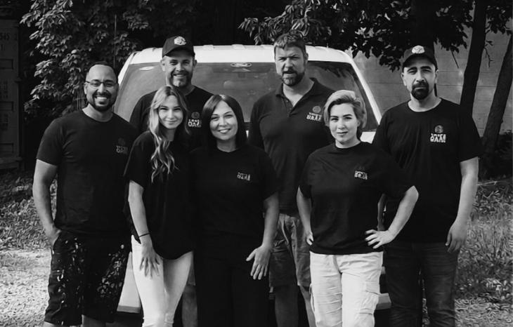 Group of seven people standing outdoors in front of a vehicle, smiling at the camera, wearing matching T-shirts.