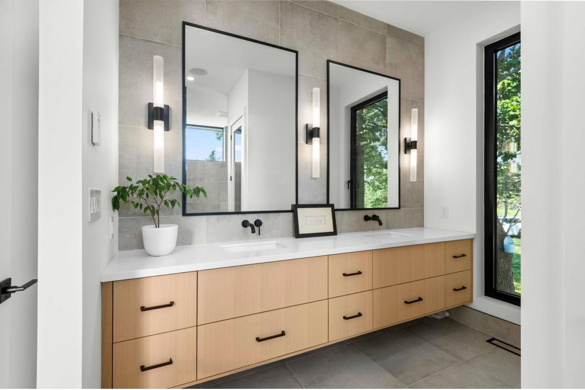 Modern bathroom vanity with wood cabinets, white countertop, dual rectangular mirrors, black wall-mounted light fixtures, a small potted plant, and a large window showing green trees outside.