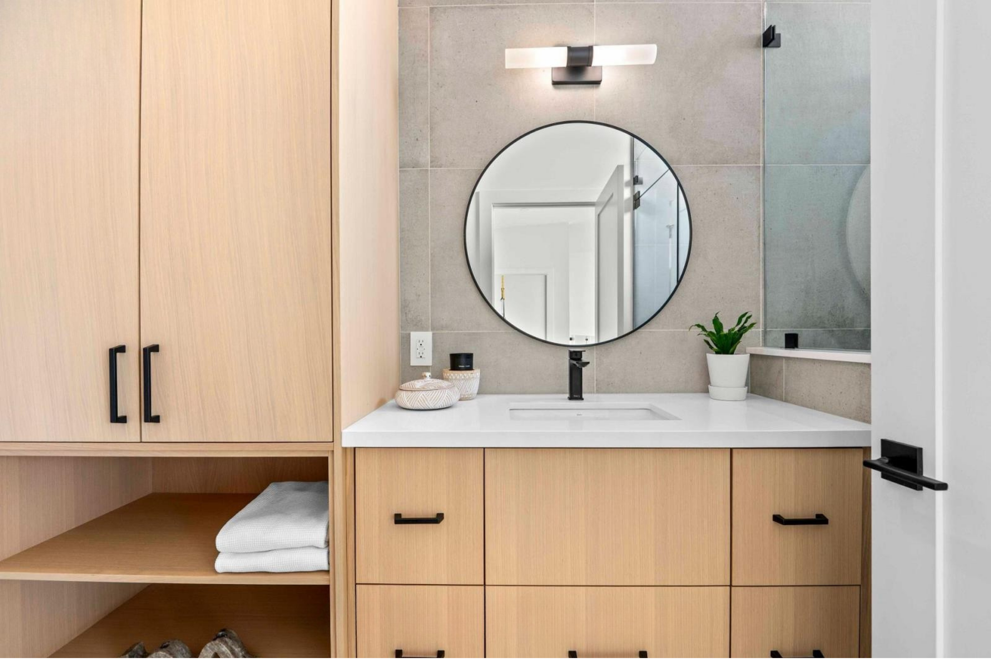 Bathroom vanity with light wood cabinets, a white countertop, a round mirror, a black faucet, and a potted plant next to the mirror.