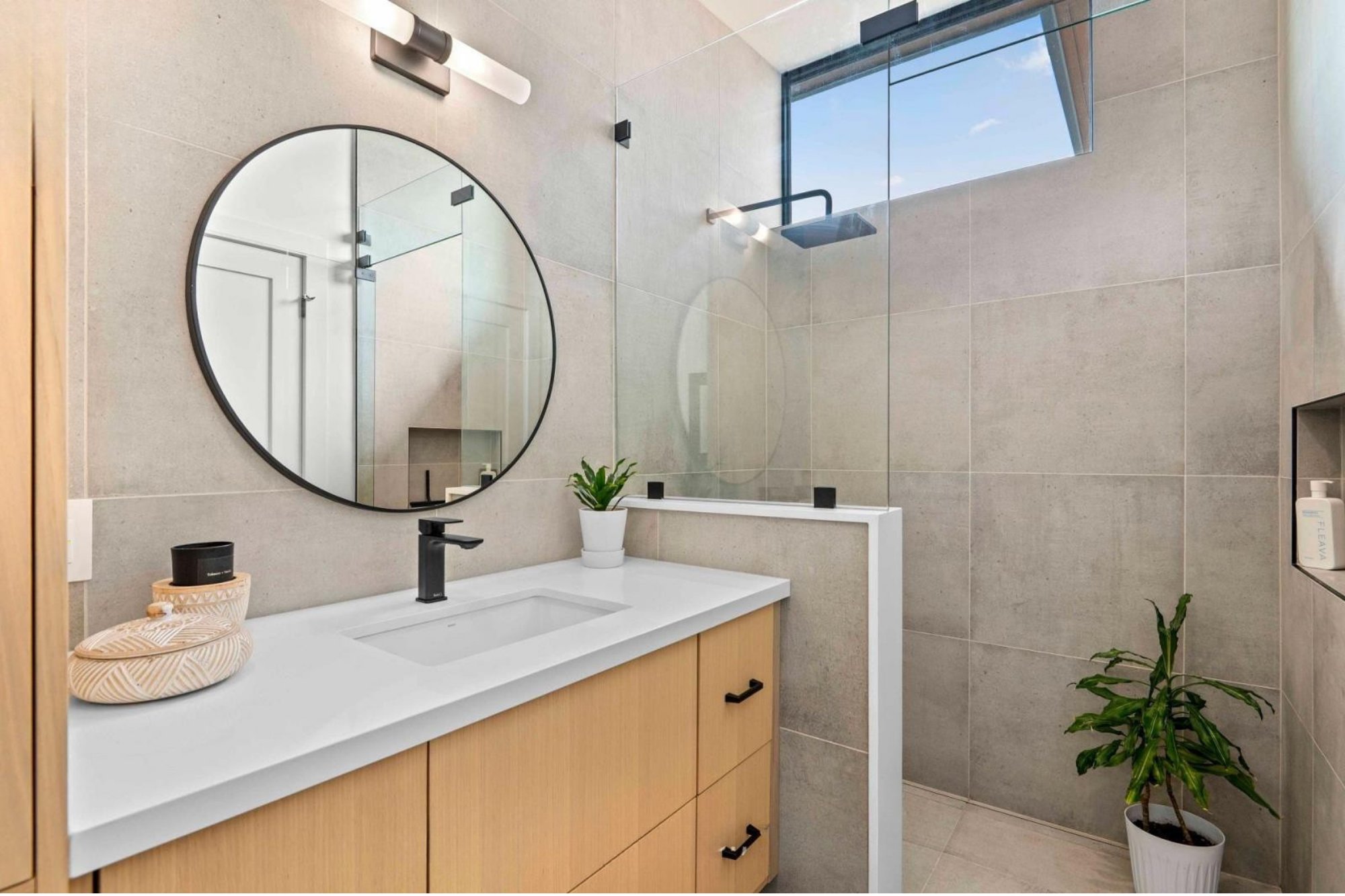 Modern bathroom with a large round mirror, black fixtures, a white sink with a wooden cabinet, a potted plant, and a glass-enclosed shower with a window showing a blue sky.