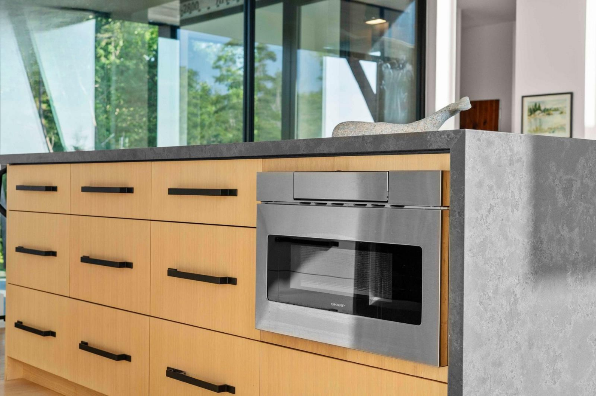 Modern kitchen with wooden cabinets, stainless steel microwave, and a stone countertop with a decorative object on it. Large windows revealing green trees outside.