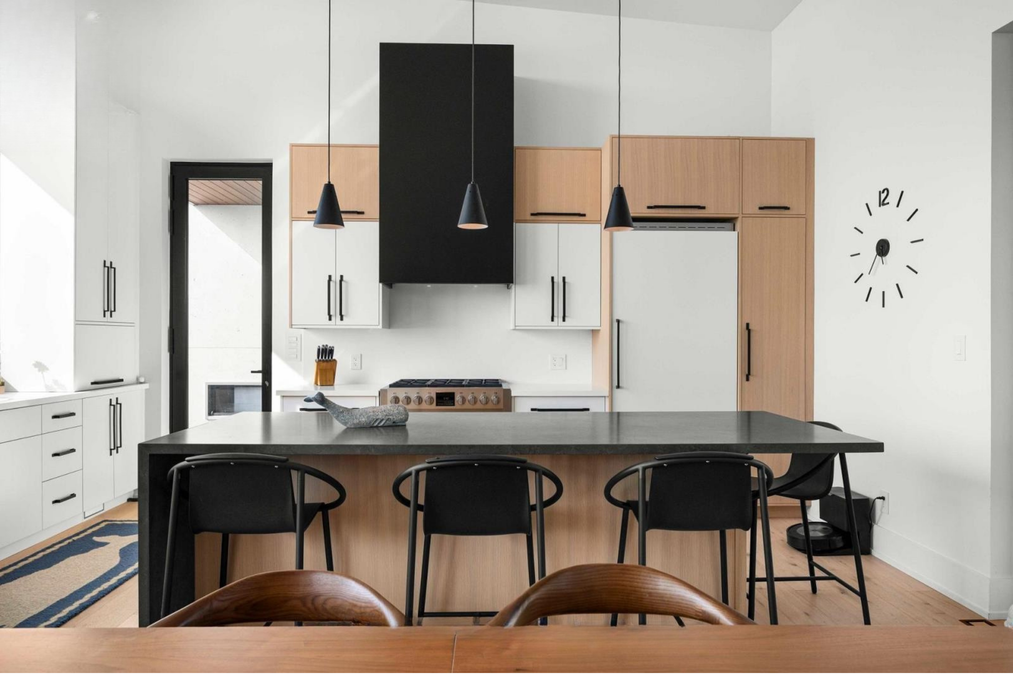 Modern kitchen with white and wood cabinets, black pendant lights, a clock on the wall, a black and beige rug, and a dark countertop with chairs.