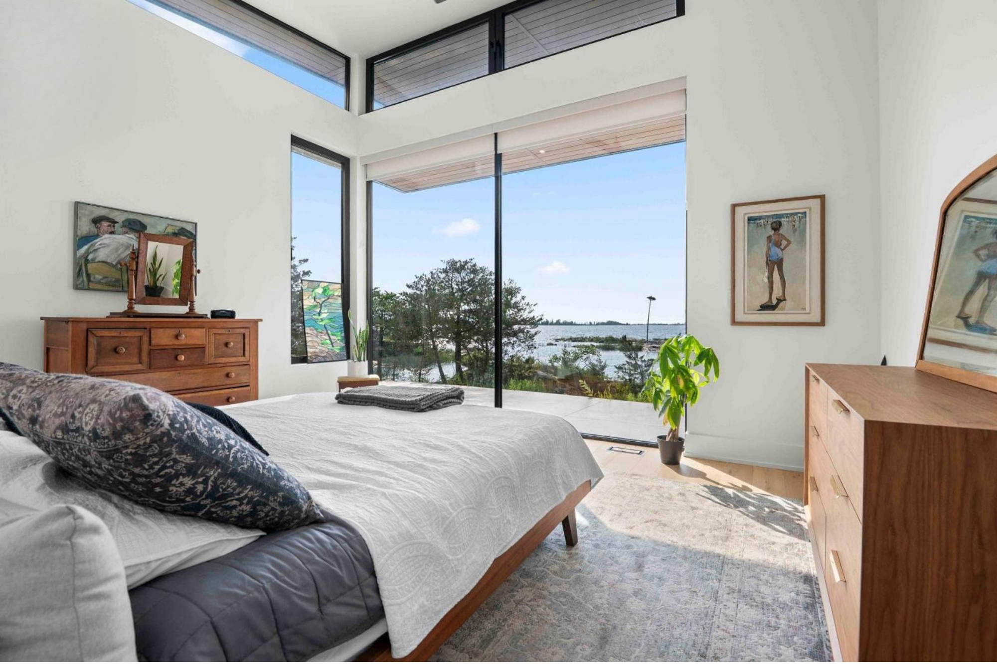 A bedroom with large floor-to-ceiling windows showing trees and a body of water outside. The bed has a white quilt and patterned pillows. There are paintings on the walls and a wooden dresser with a mirror, some plants, and artwork nearby.