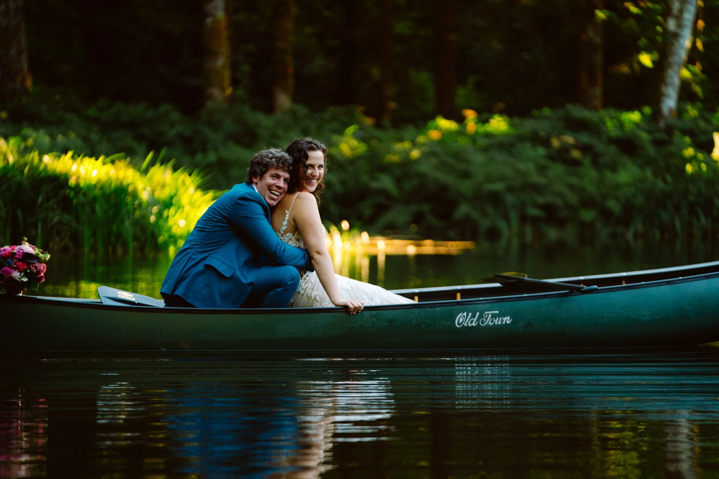 A joyful couple in wedding attire sitting on a boat named 'Old Town' on a calm river surrounded by lush greenery, with sunlight filtering through trees in the background.