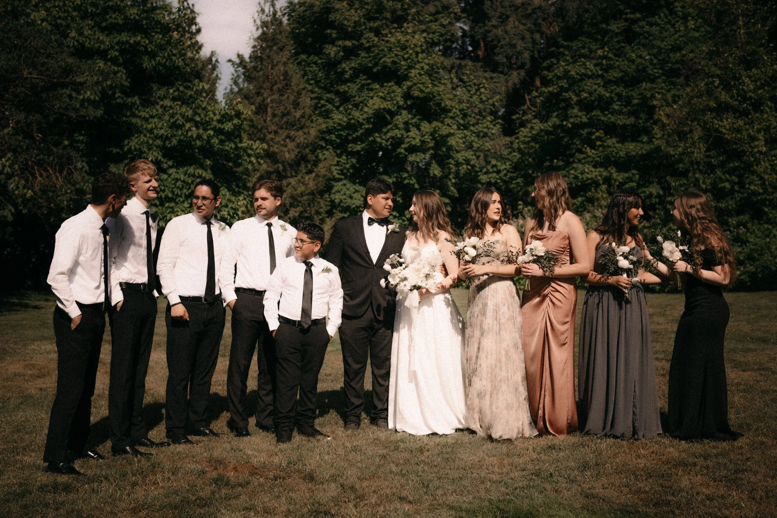 A group of people dressed in formal attire standing outdoors in front of trees, likely at a wedding or similar celebration.