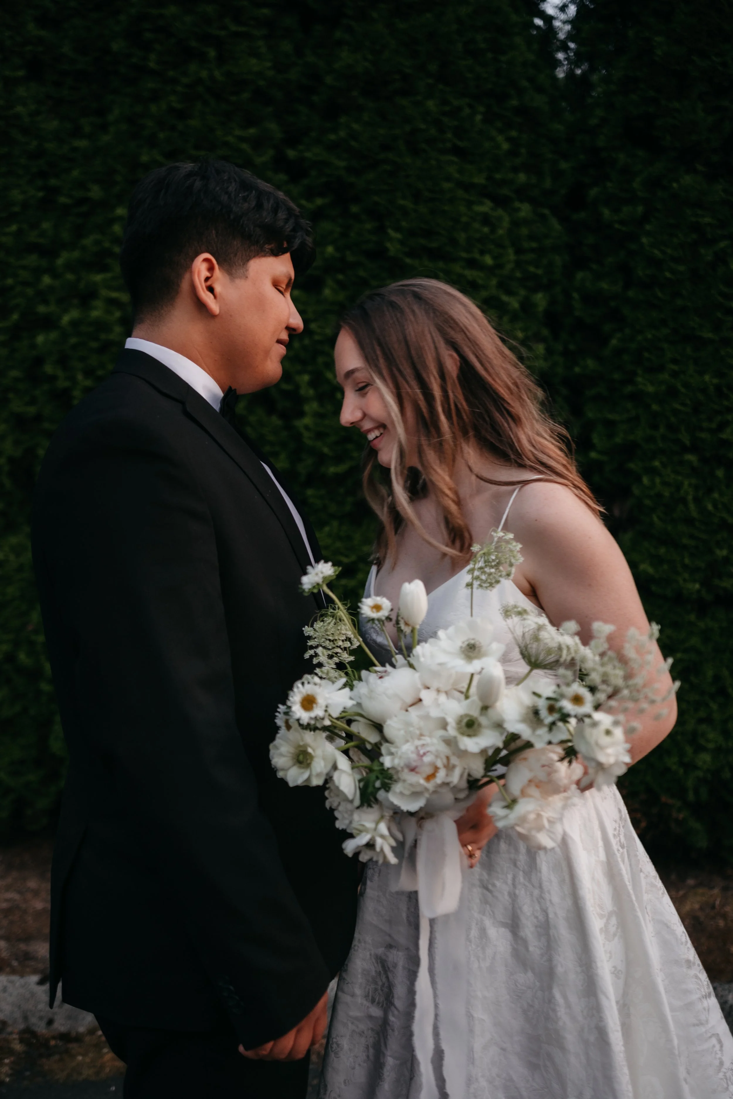 A couple dressed in wedding attire holding hands and smiling, with the bride holding a large bouquet of white flowers, standing outdoors against a green hedge background.