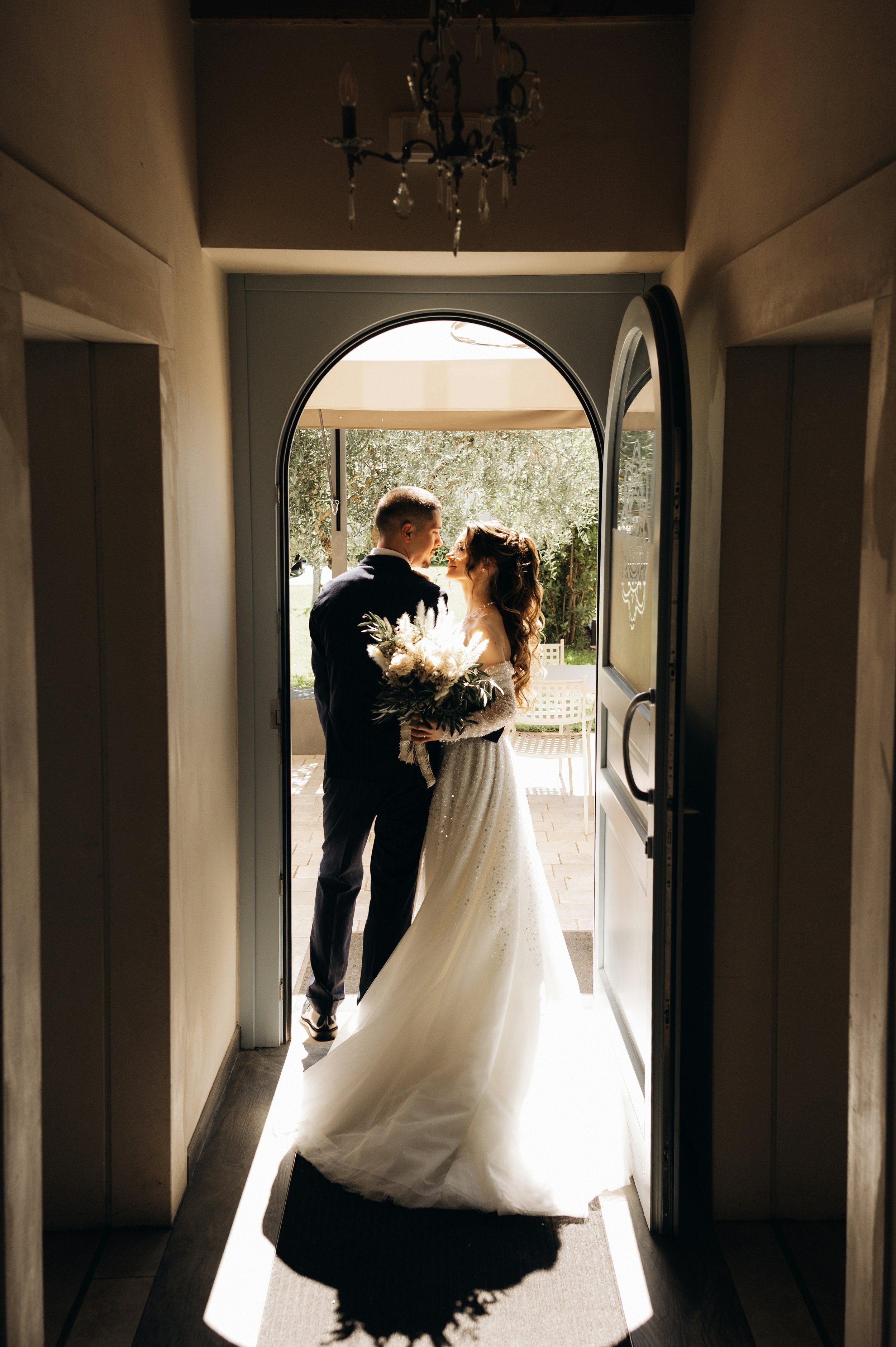 A bride and groom standing together in a doorway, sharing an intimate moment, with sunlight shining on them and trees visible in the background.