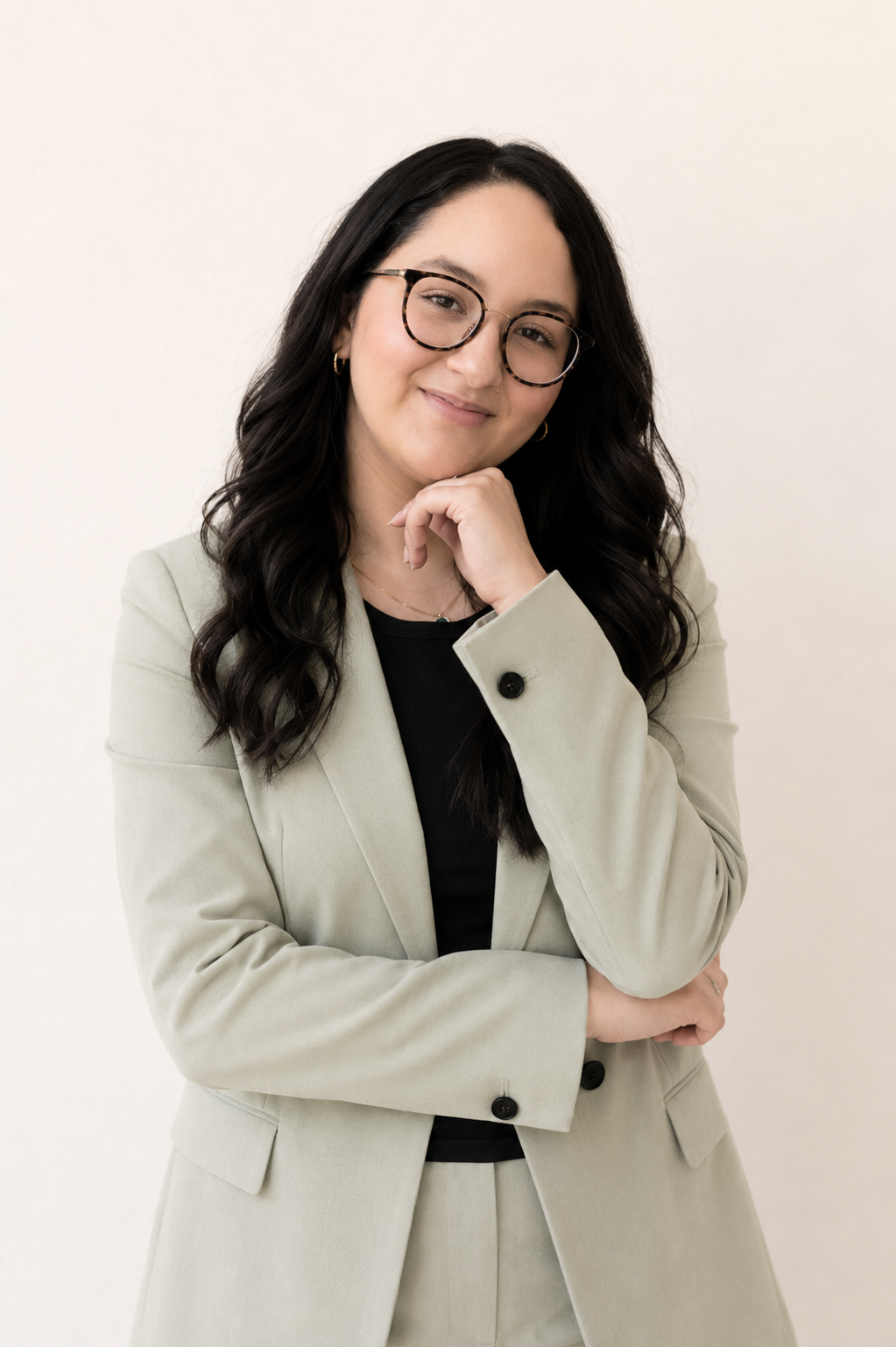 A woman with long dark hair, glasses, and earrings posing confidently in a beige suit with a black top against a plain white background.
