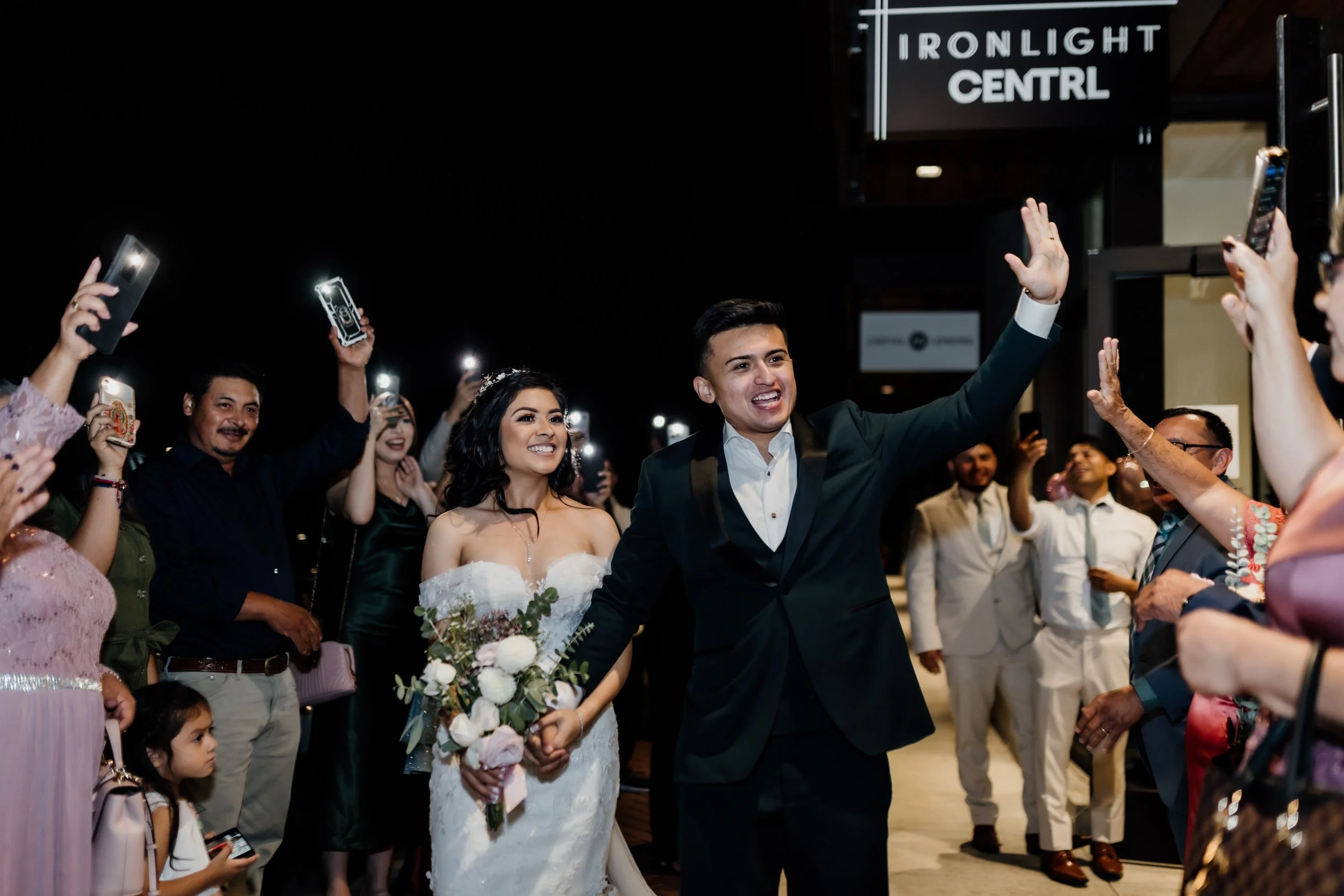 A bride and groom at their wedding reception, smiling and waving at guests. The bride is holding a bouquet of flowers, and guests are taking photos with their phones.