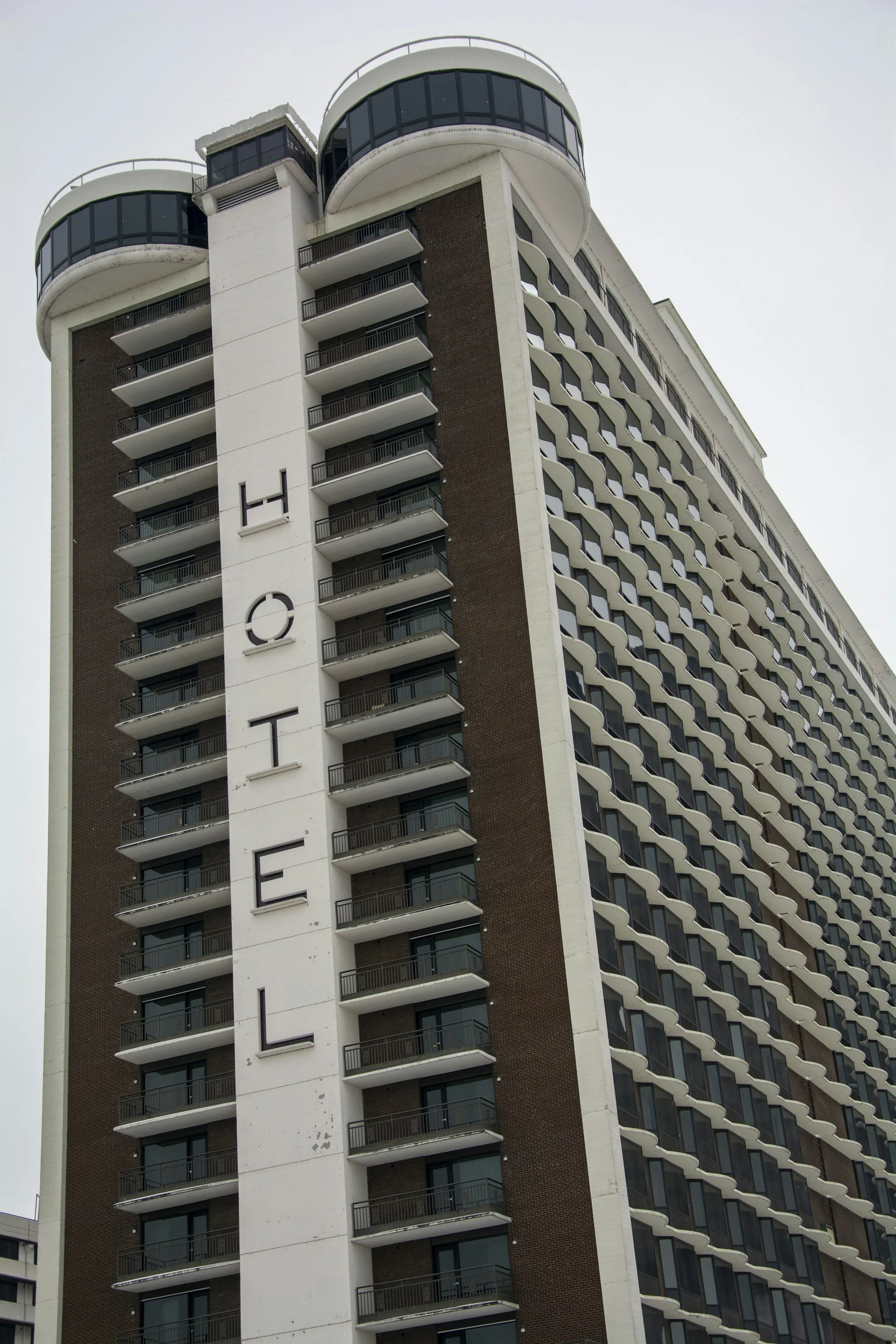 A tall modern high-rise building with numerous curved balconies and a large vertical sign spelling out 'HOTEL' on the front