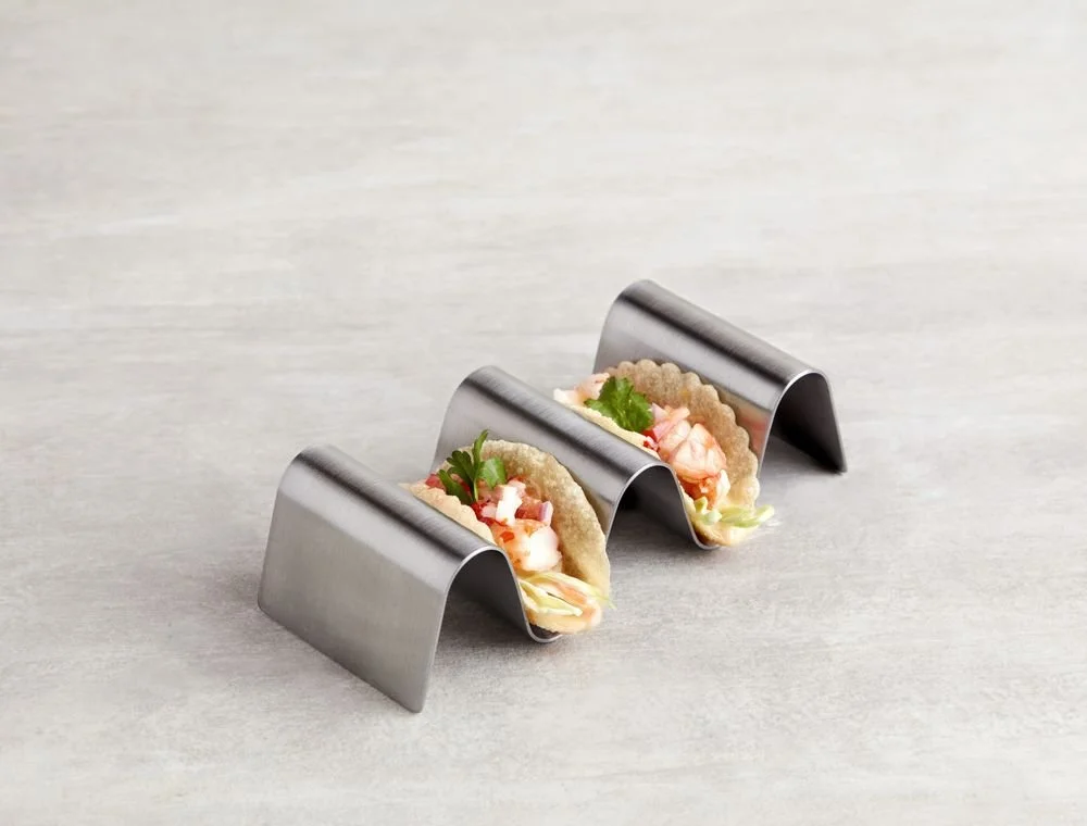 Two small seafood tacos in metal holders on a light-colored surface.