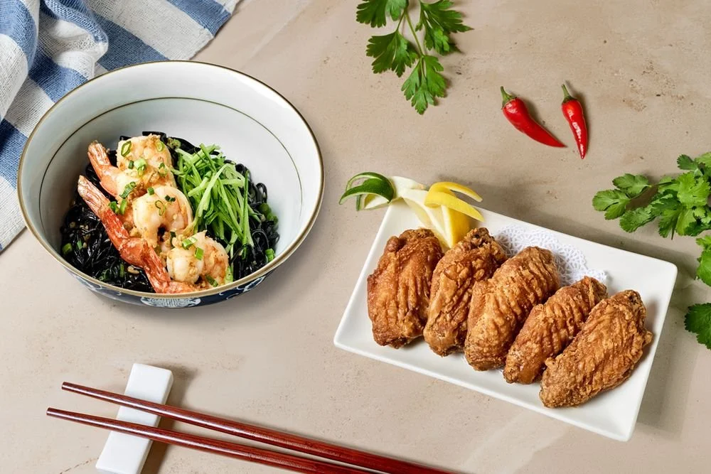 A bowl of shrimp and vegetable pasta and a plate of fried chicken tenders with lemon wedges, placed on a light-colored surface with garnishing herbs and red chili peppers nearby.