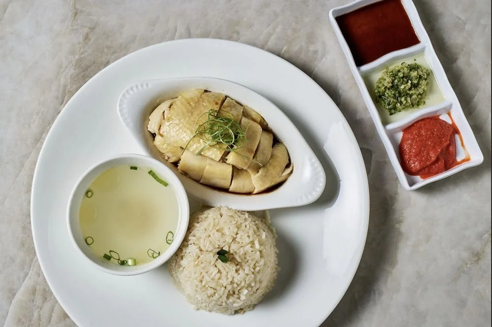 Hainanese chicken rice served on a white plate with steamed chicken, rice, chicken broth, and dipping sauces, with a side of three additional sauces in a divided dish.