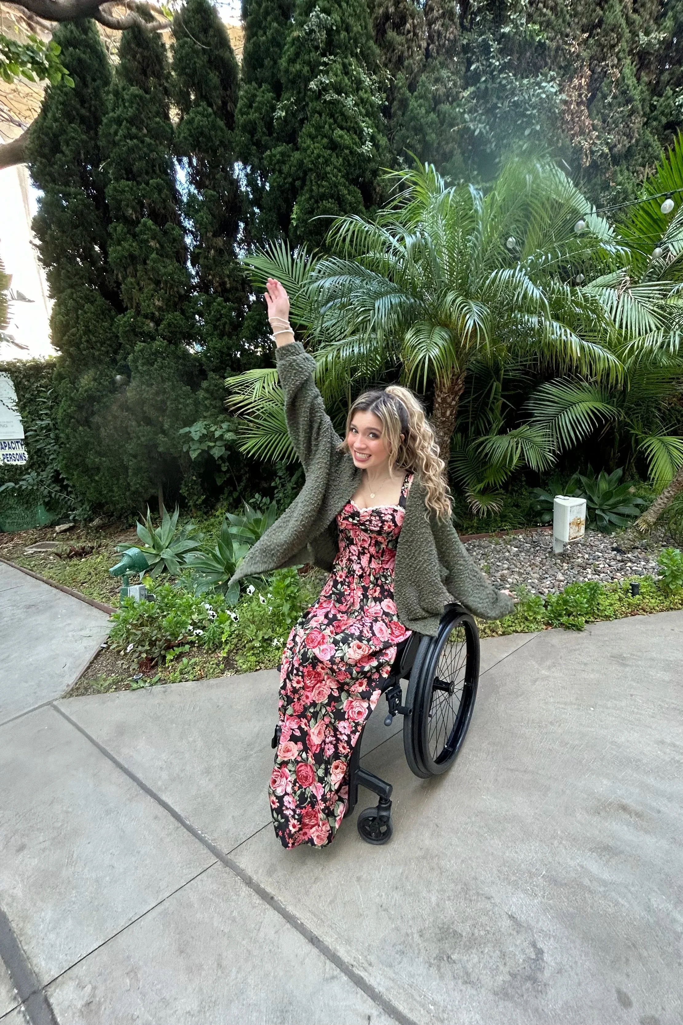 A smiling woman with curly blonde hair wearing a floral dress and gray cardigan, sitting in a wheelchair outdoors with lush green trees and plants in the background.