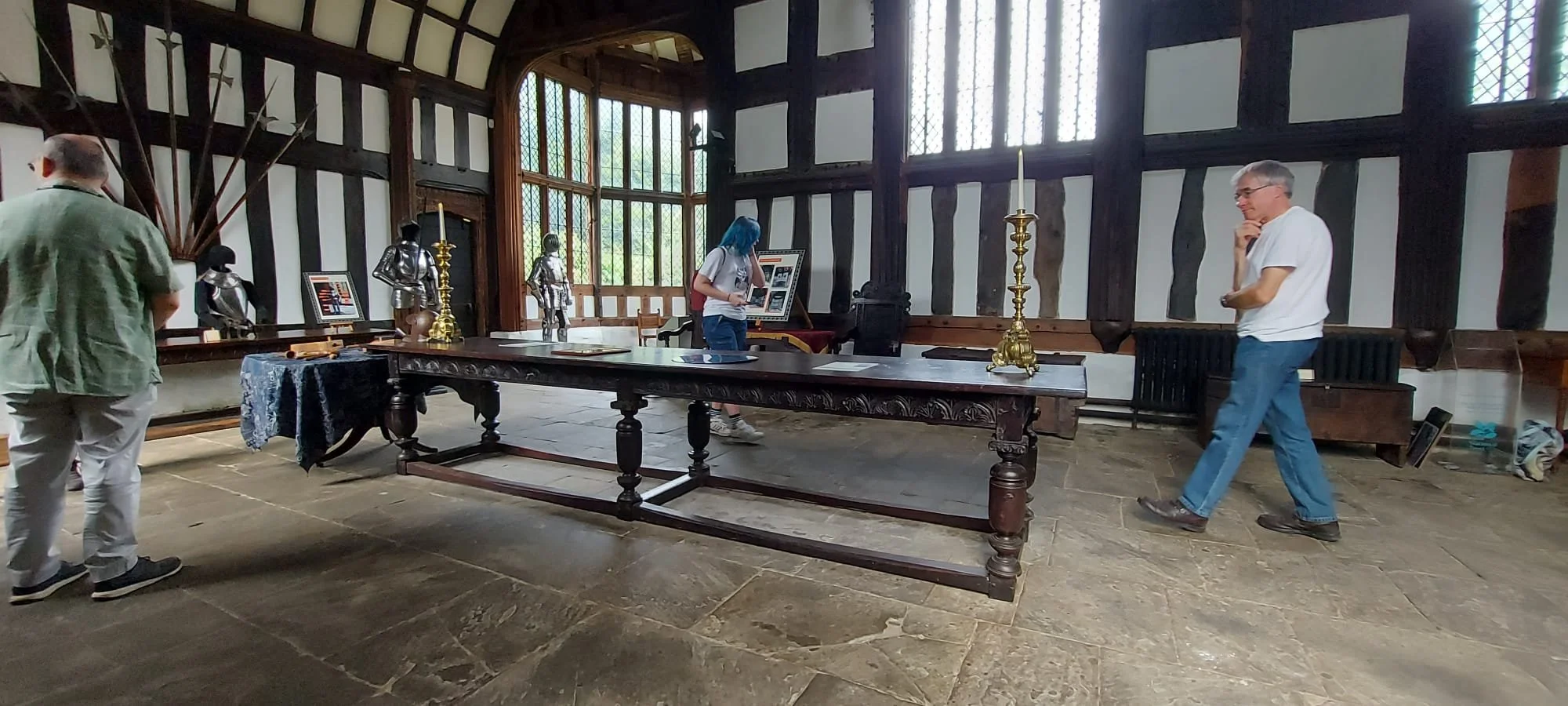 A Tudor Great Hall interior, featuring a large wooden table with candles, medieval armour, and swords on display. There is a person observing the displays, with tall windows letting in natural light, and the walls have dark wooden beams.