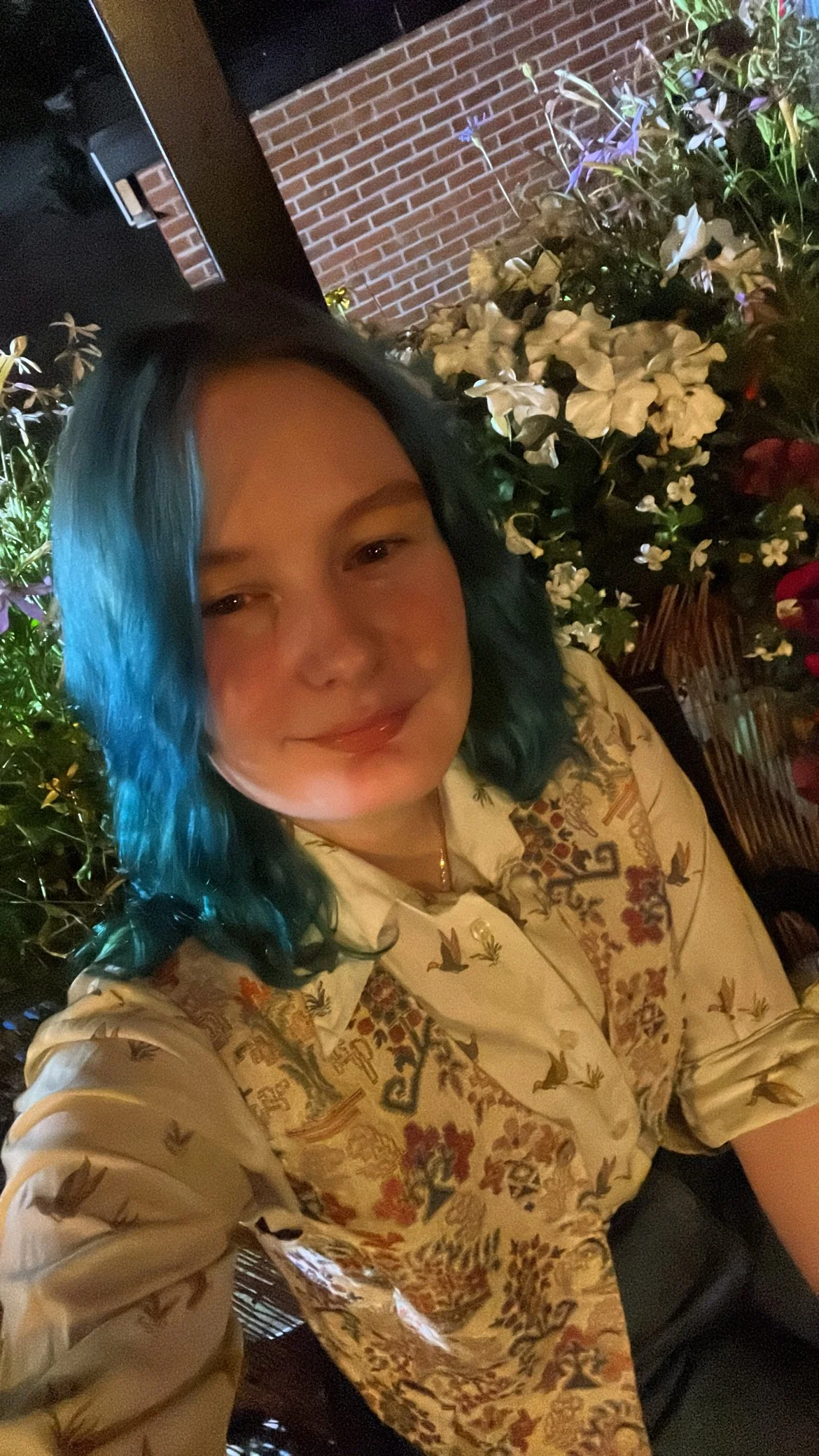 A man with blue hair taking a selfie in front of flowers against a brick wall at night.
