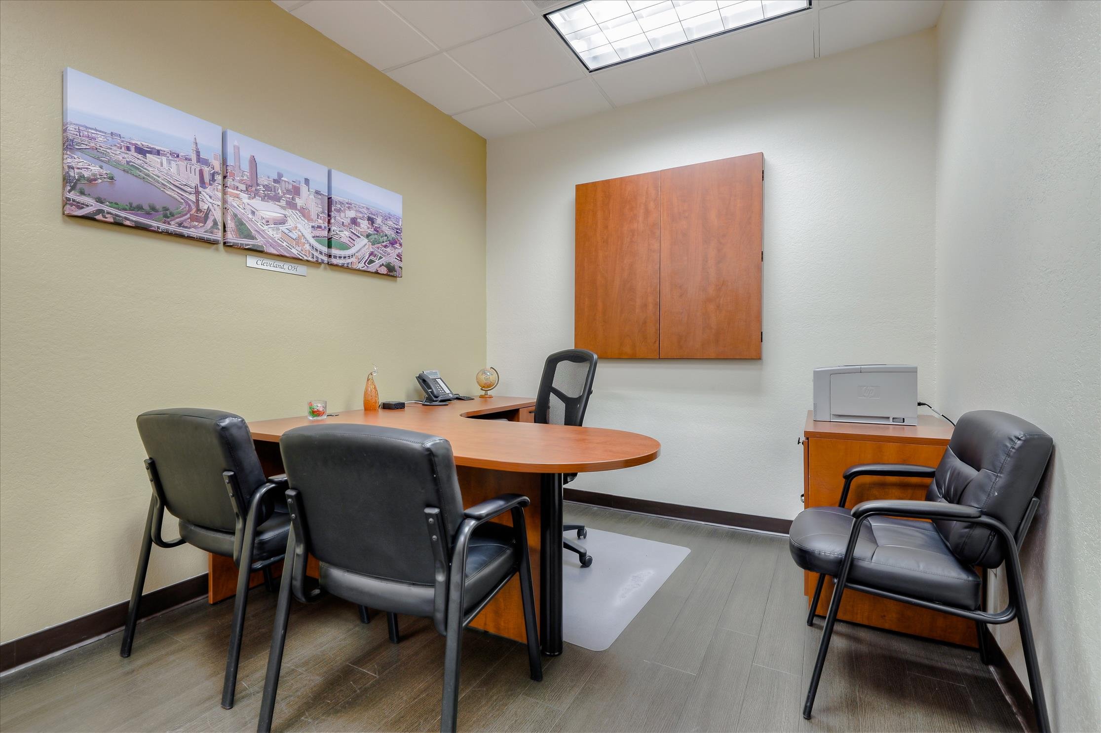 An office conference room with a wooden table surrounded by black leather chairs. On the wall, there is a large cityscape photograph of Cleveland, Ohio, and a wooden cabinet is mounted on the wall. A desk in the corner has a phone, a small globe, and a decorative item. The ceiling has a fluorescent light panel, and the floor has a dark wood or laminate finish.