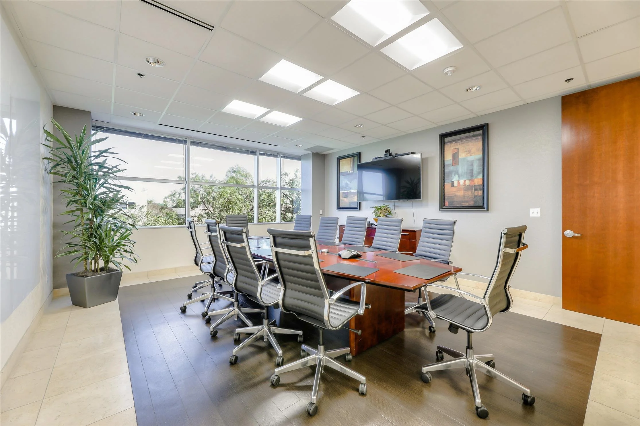 A modern conference room with a long wooden table surrounded by gray office chairs, large windows with a view of trees outside, a wall-mounted television, and artwork on the walls.
