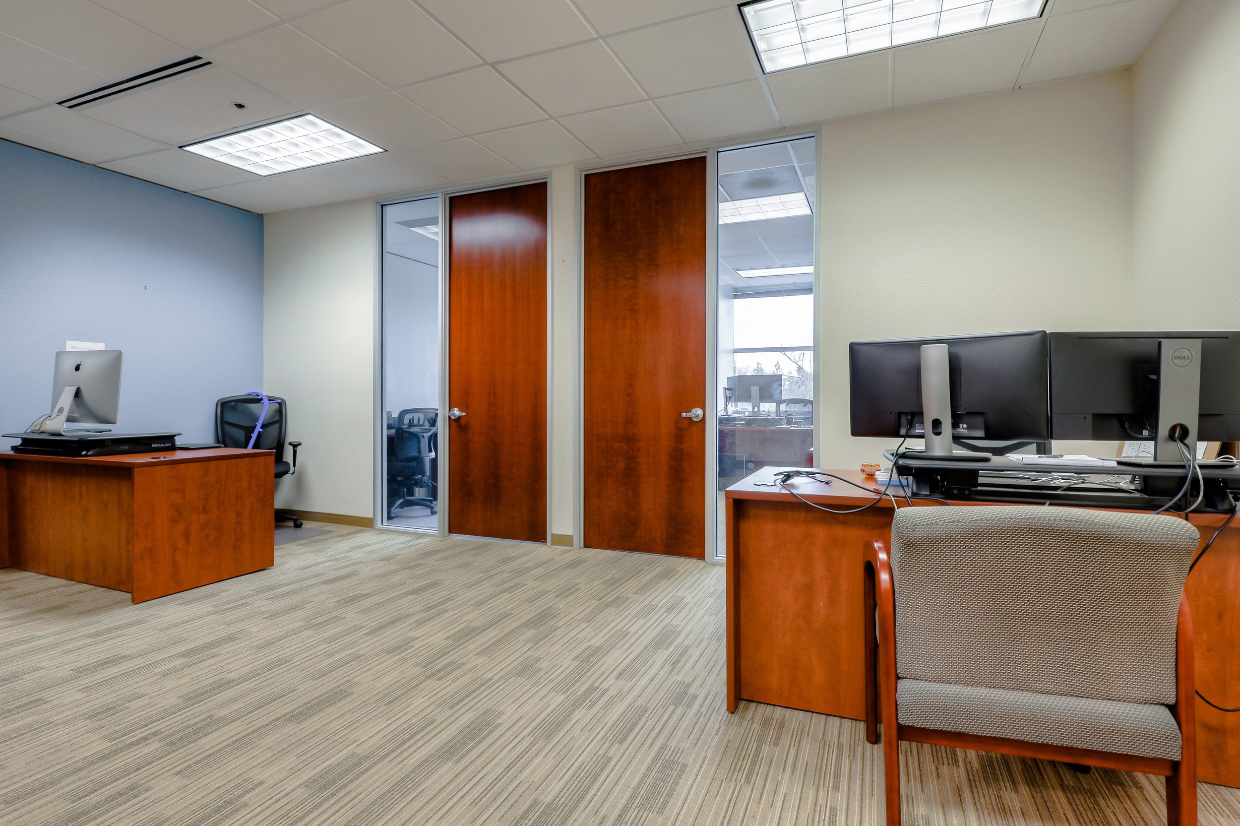 An office with two desks, one with a desktop computer and chair, and the other with dual monitors, both made of wood with a red-brown finish. There are glass doors leading to a conference room or hallway, windows with blinds, and beige carpet flooring.